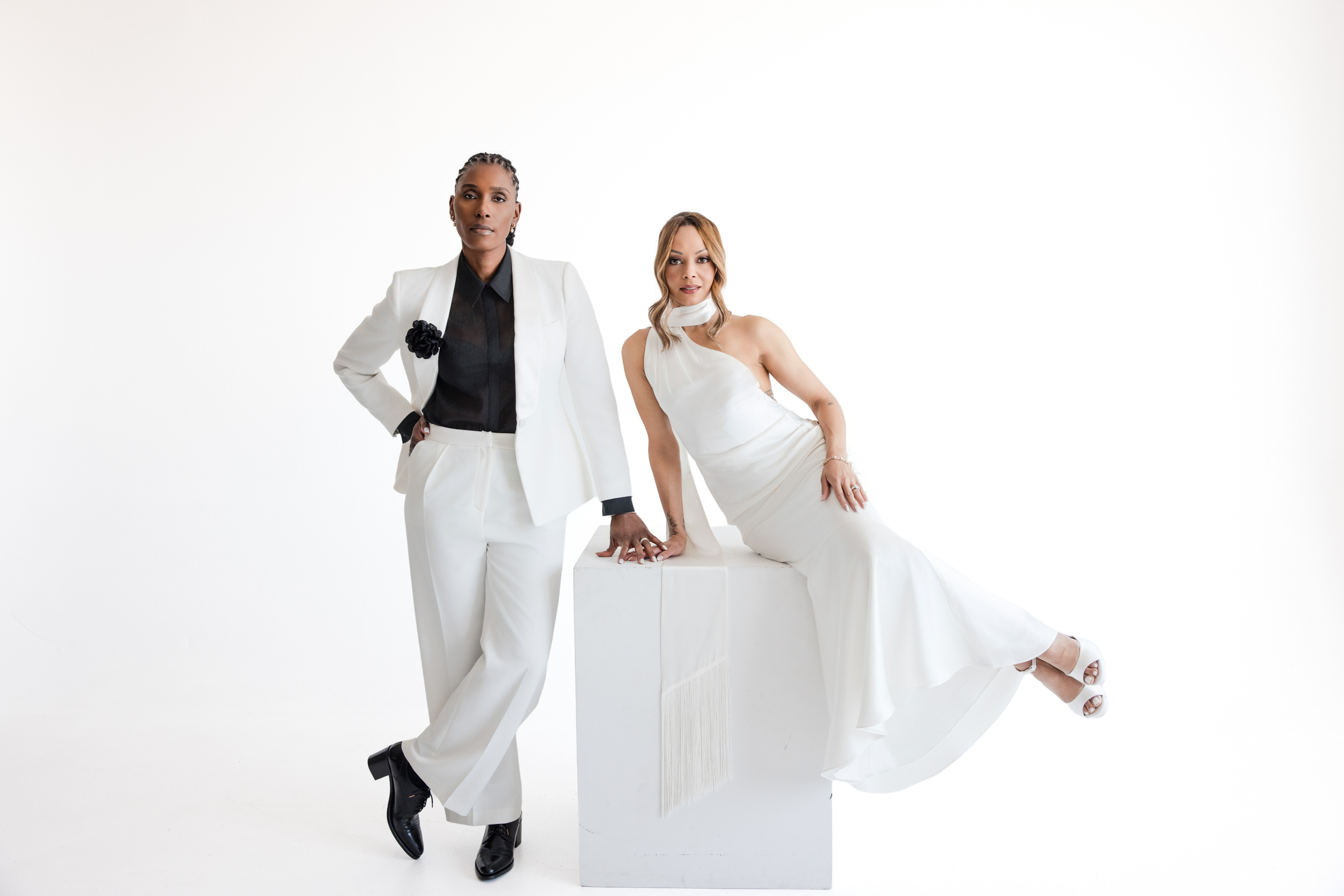Two people in formal white outfits pose against a white background during their NYC studio engagement session; one stands in a sleek white suit, while the other sits elegantly on a white cube in a flowing white dress.