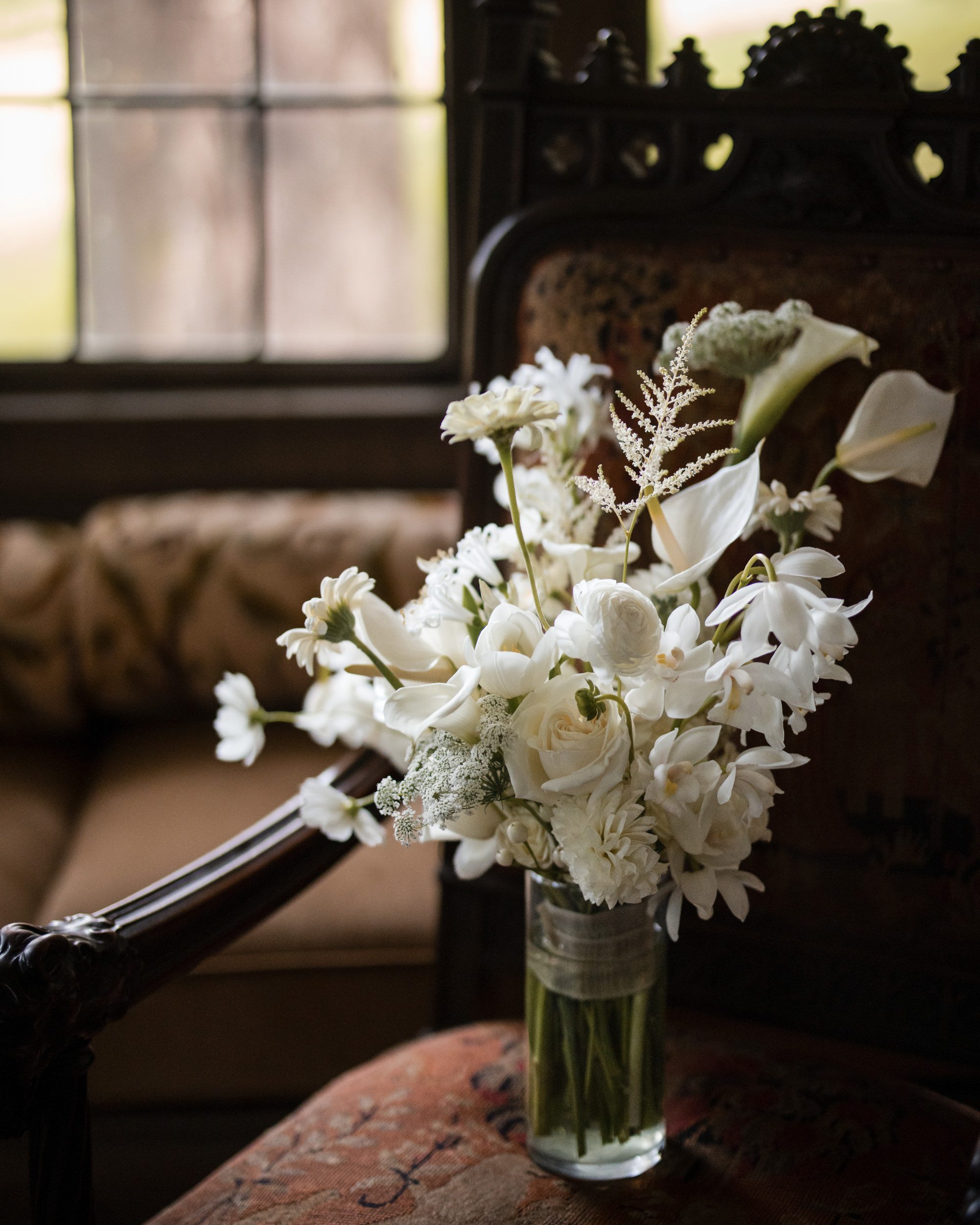 A glass vase with an arrangement of white flowers sits on an ornate, upholstered chair near a window, evoking the timeless elegance of a Lyndhurst Mansion wedding.