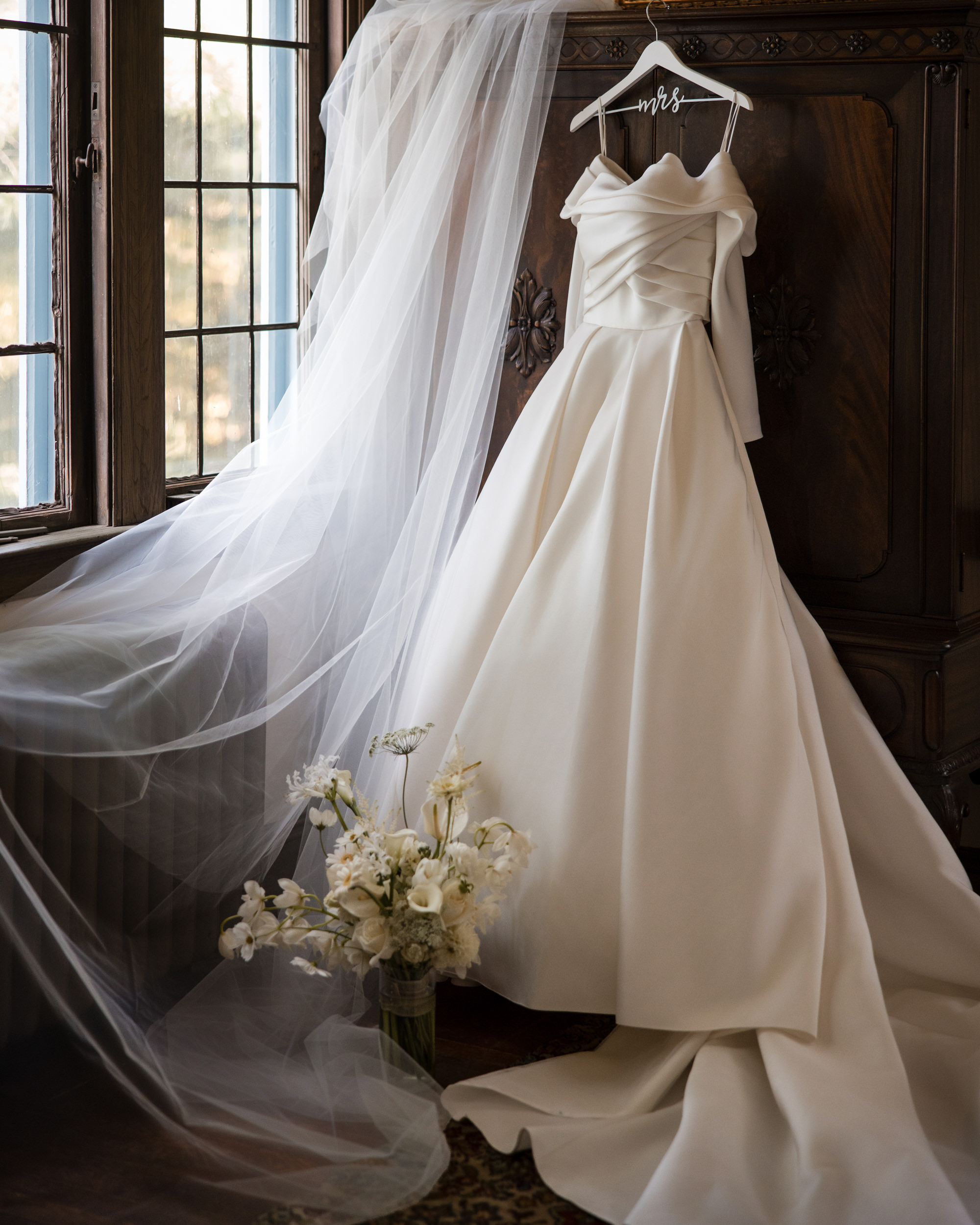 An off-the-shoulder white wedding dress hangs on a wooden wardrobe at a lyndhurst mansion wedding, with a long veil and a bouquet of white flowers nearby.