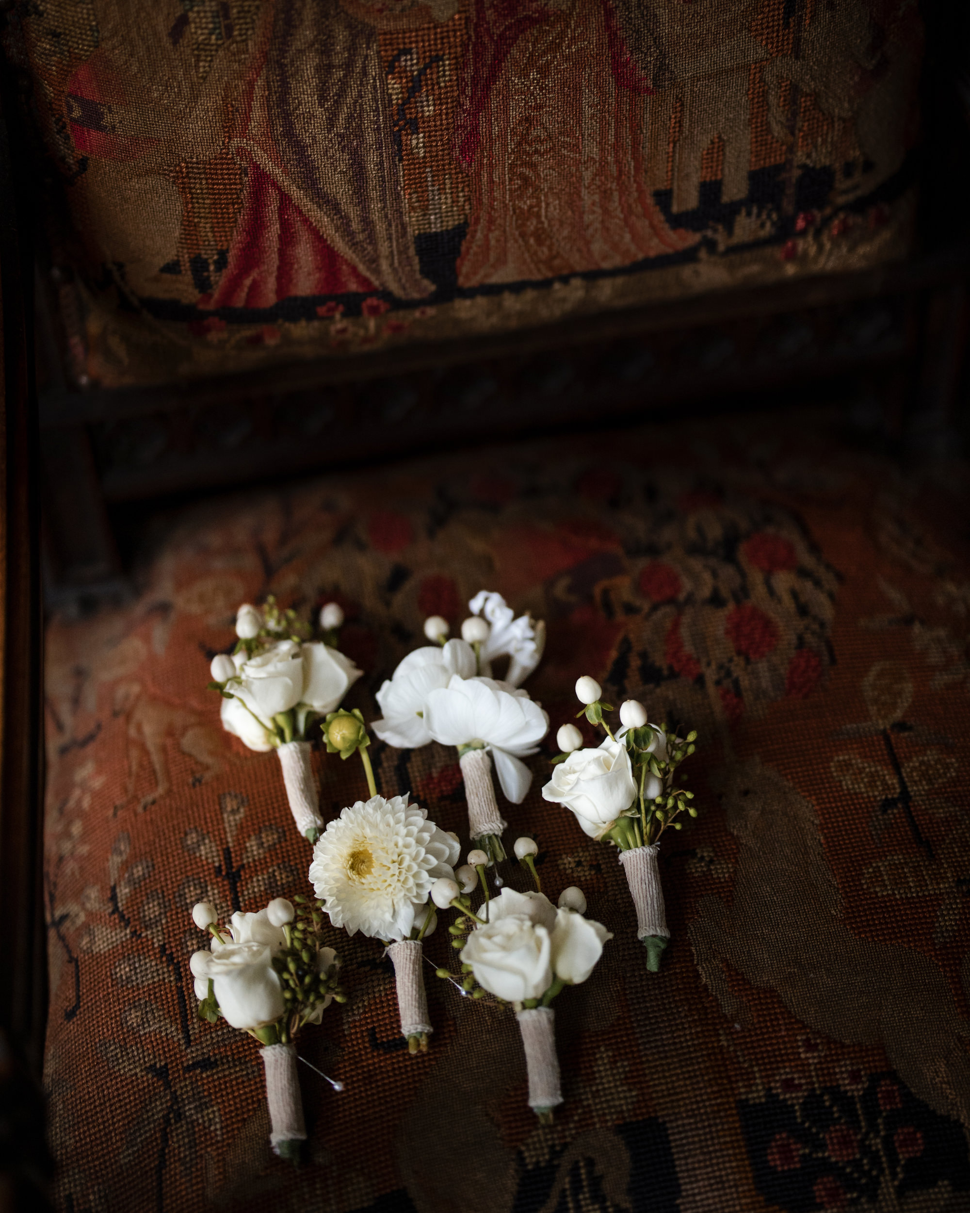 Six small white flower boutonnieres with wrapped stems are arranged on a vintage, tapestry-upholstered chair with a detailed woven pattern, capturing the timeless elegance perfect for a Lyndhurst Mansion wedding.