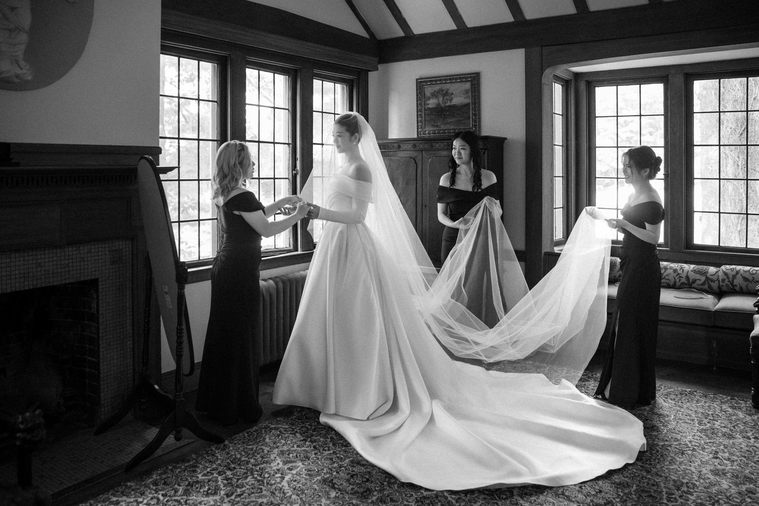 A bride in a wedding gown stands indoors at her Lyndhurst Mansion wedding as three bridesmaids in dark dresses help arrange her veil and dress train.