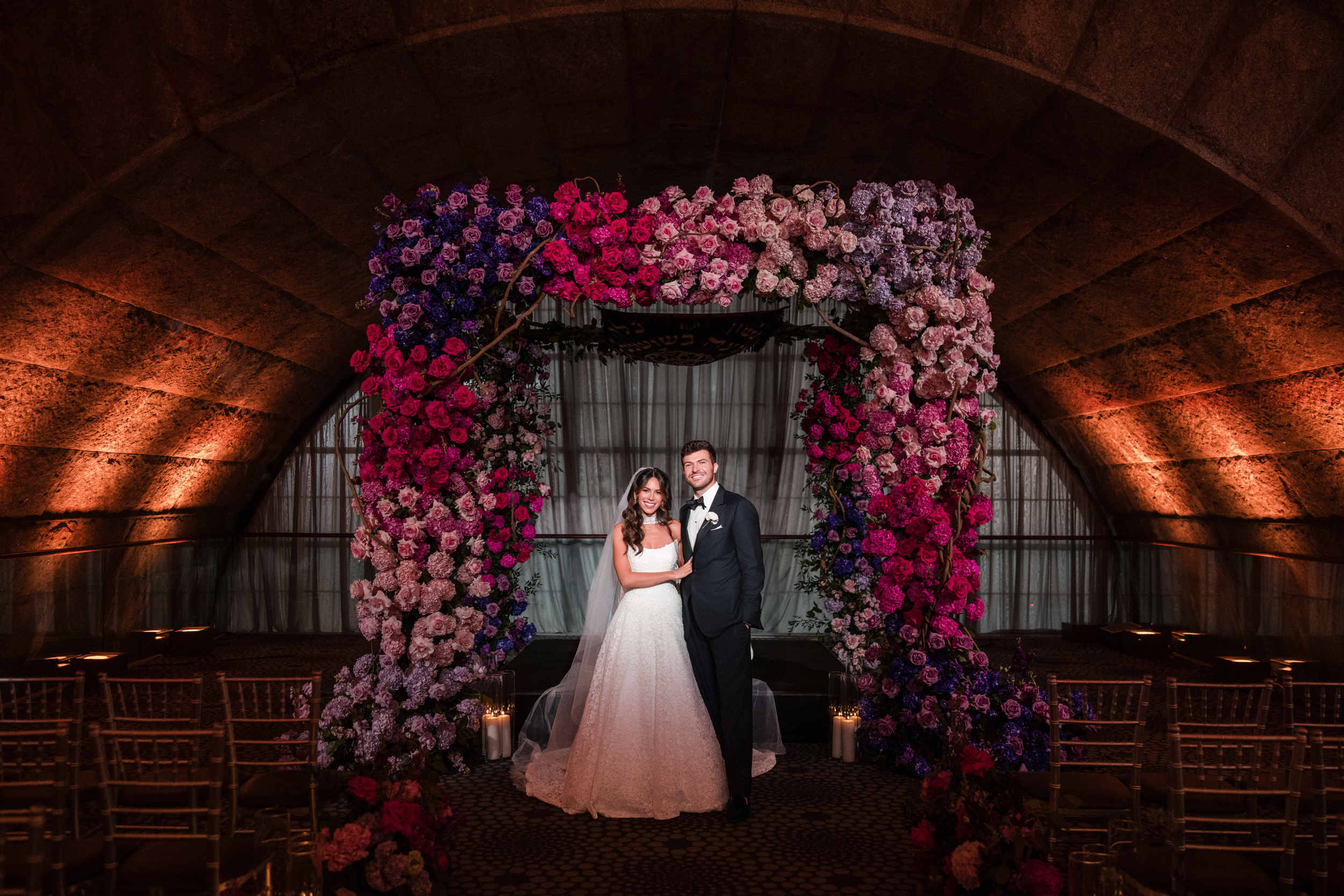 A bride and groom stand under a vibrant floral arch in a dimly lit venue at Guastavino’s wedding, with rows of empty chairs arranged in front of them.