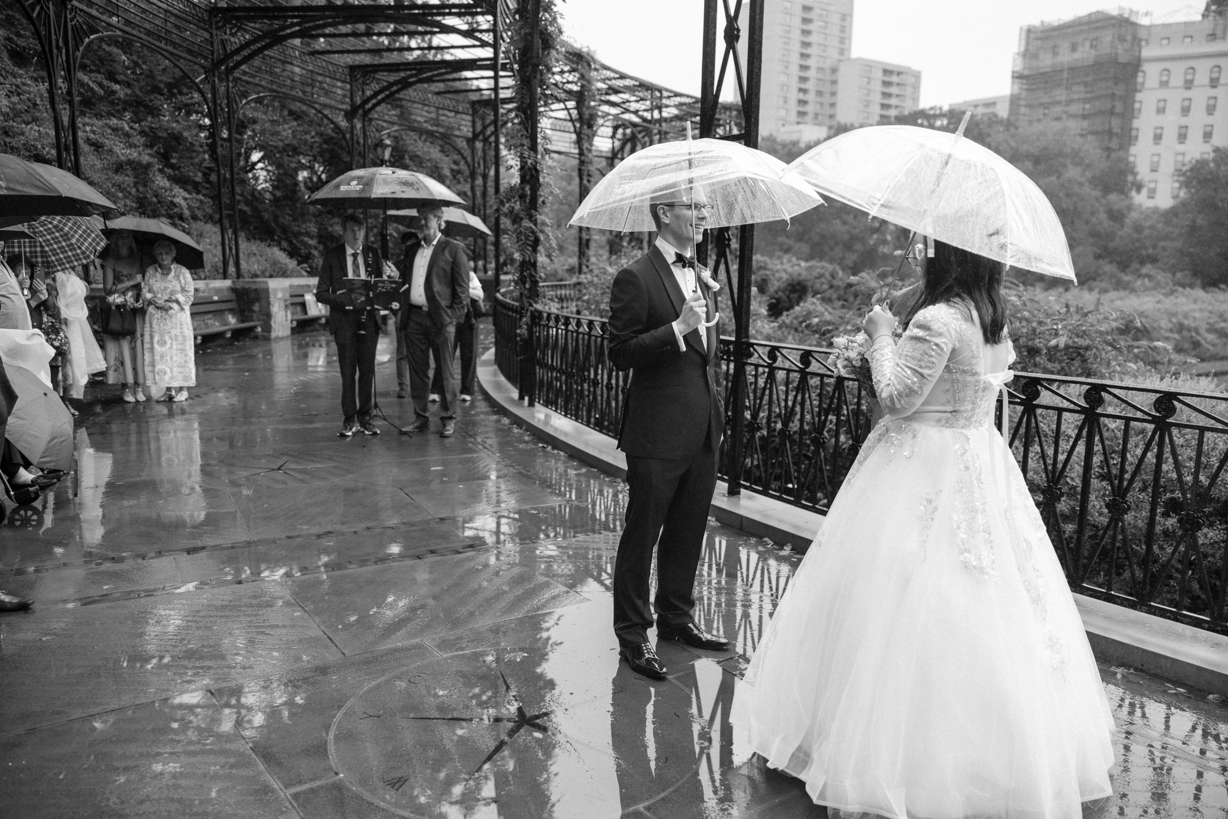 A bride and groom stand under clear umbrellas on a rainy day, facing each other during their Conservatory Garden wedding, while guests with umbrellas watch nearby on a wet, covered walkway.