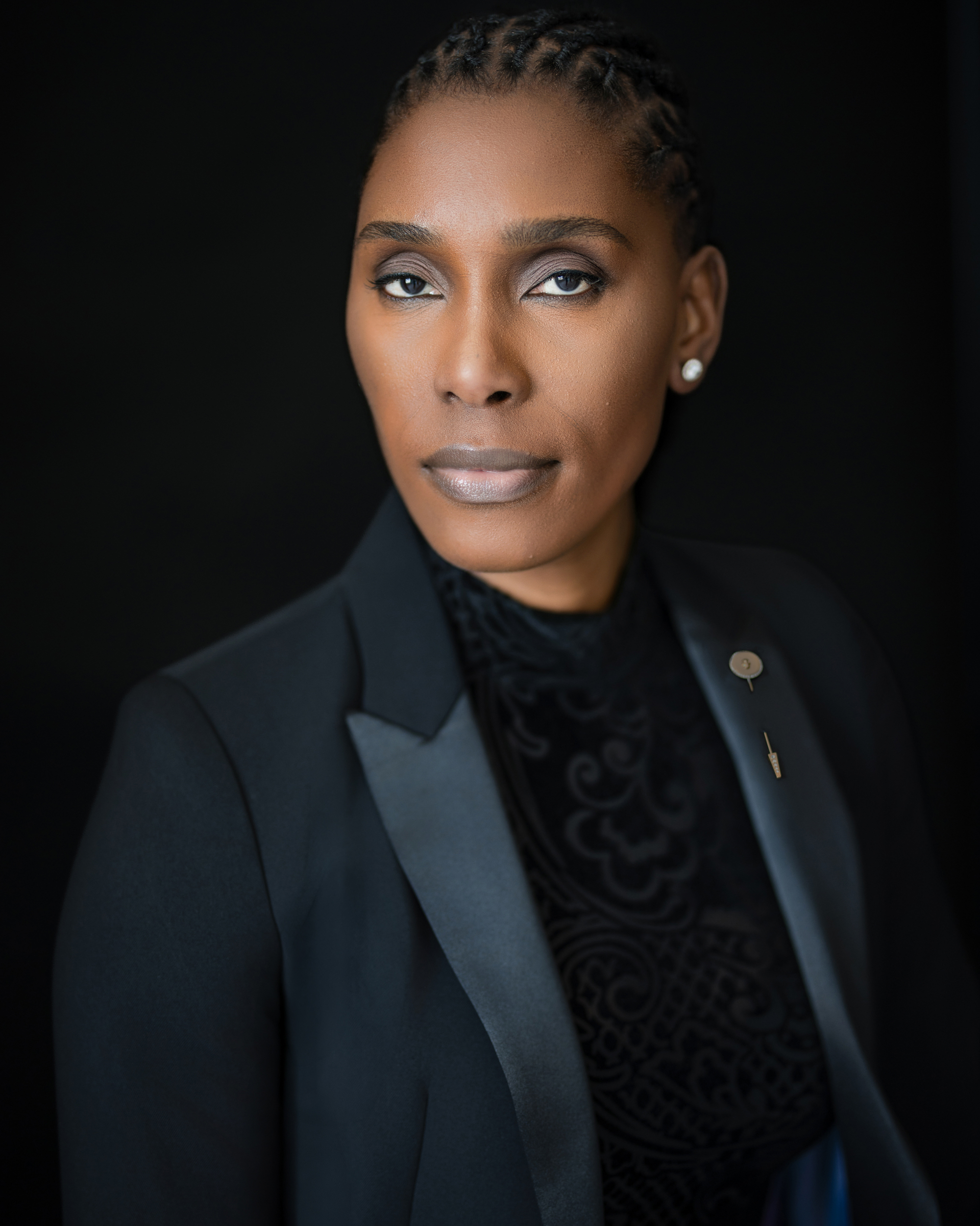 A person in a black suit jacket and patterned black top poses against a dark background, with neatly styled hair and a calm, confident expression, capturing the sophisticated vibe of an NYC studio engagement session.