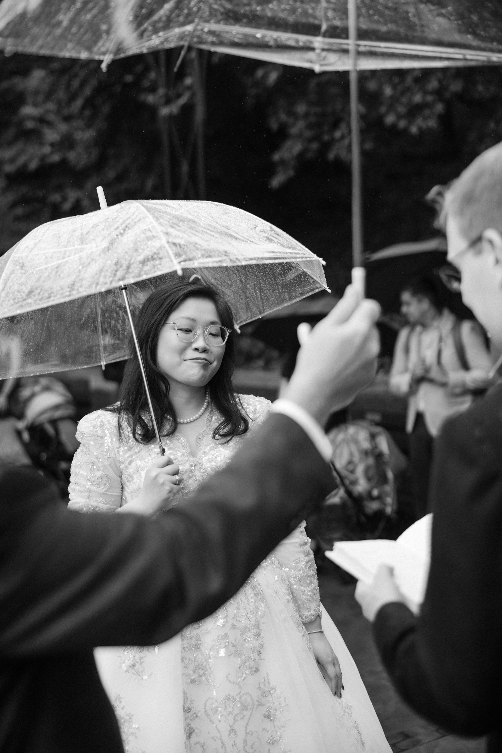 A woman in a wedding dress stands under an umbrella while someone reads from a book at an outdoor event on a rainy day, capturing the romantic atmosphere of a Conservatory Garden wedding.