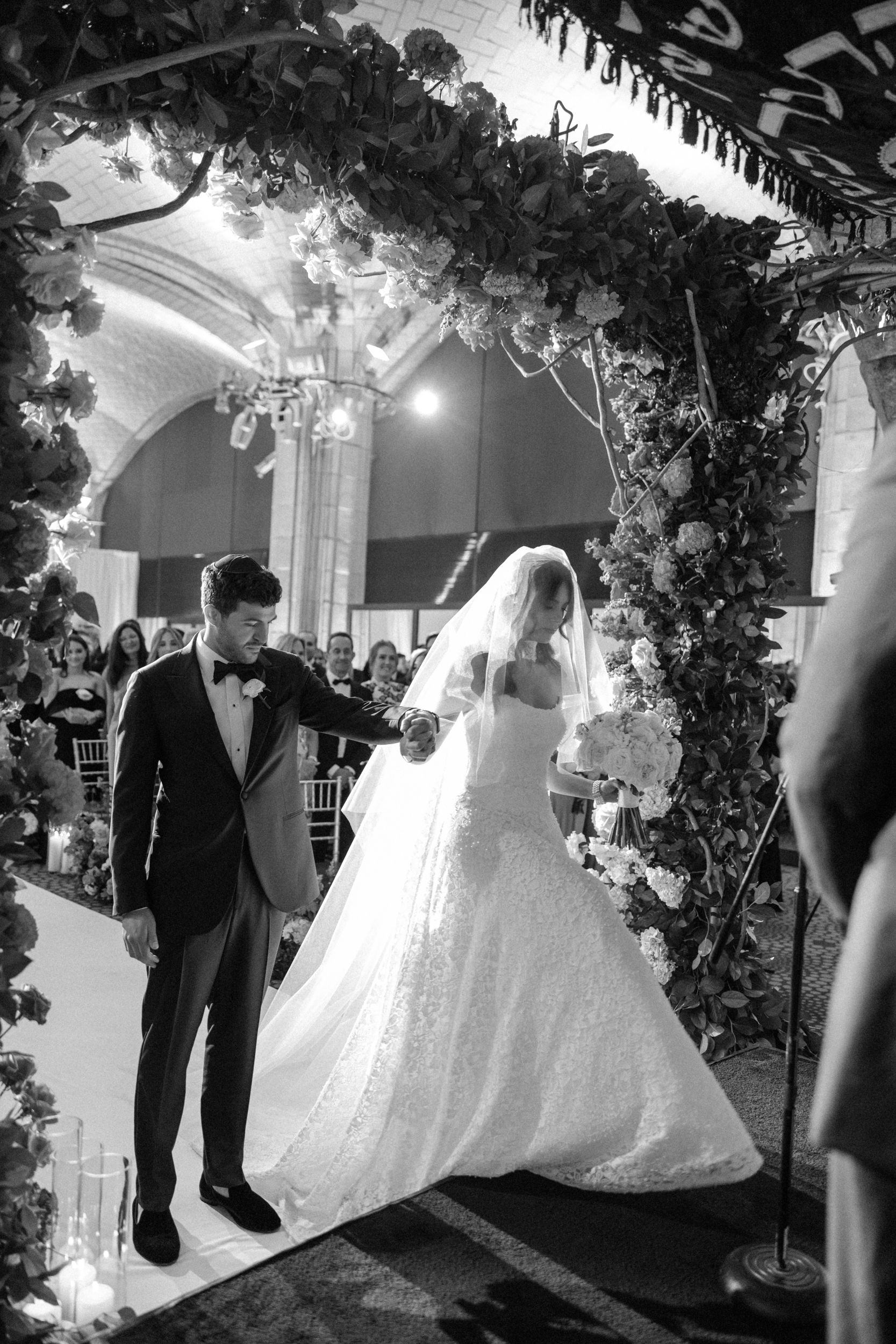 A bride and groom stand together under a floral wedding arch, surrounded by guests, in an elegantly decorated indoor venue, capturing the timeless charm of a Guastavino’s wedding.