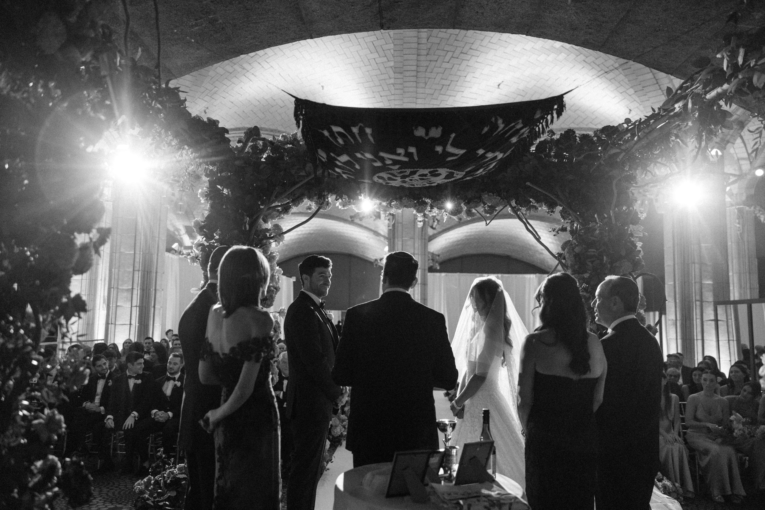 A beautiful Guastavino’s wedding ceremony unfolds under a floral chuppah, with the couple, officiant, and family members standing together while guests are seated in the background.