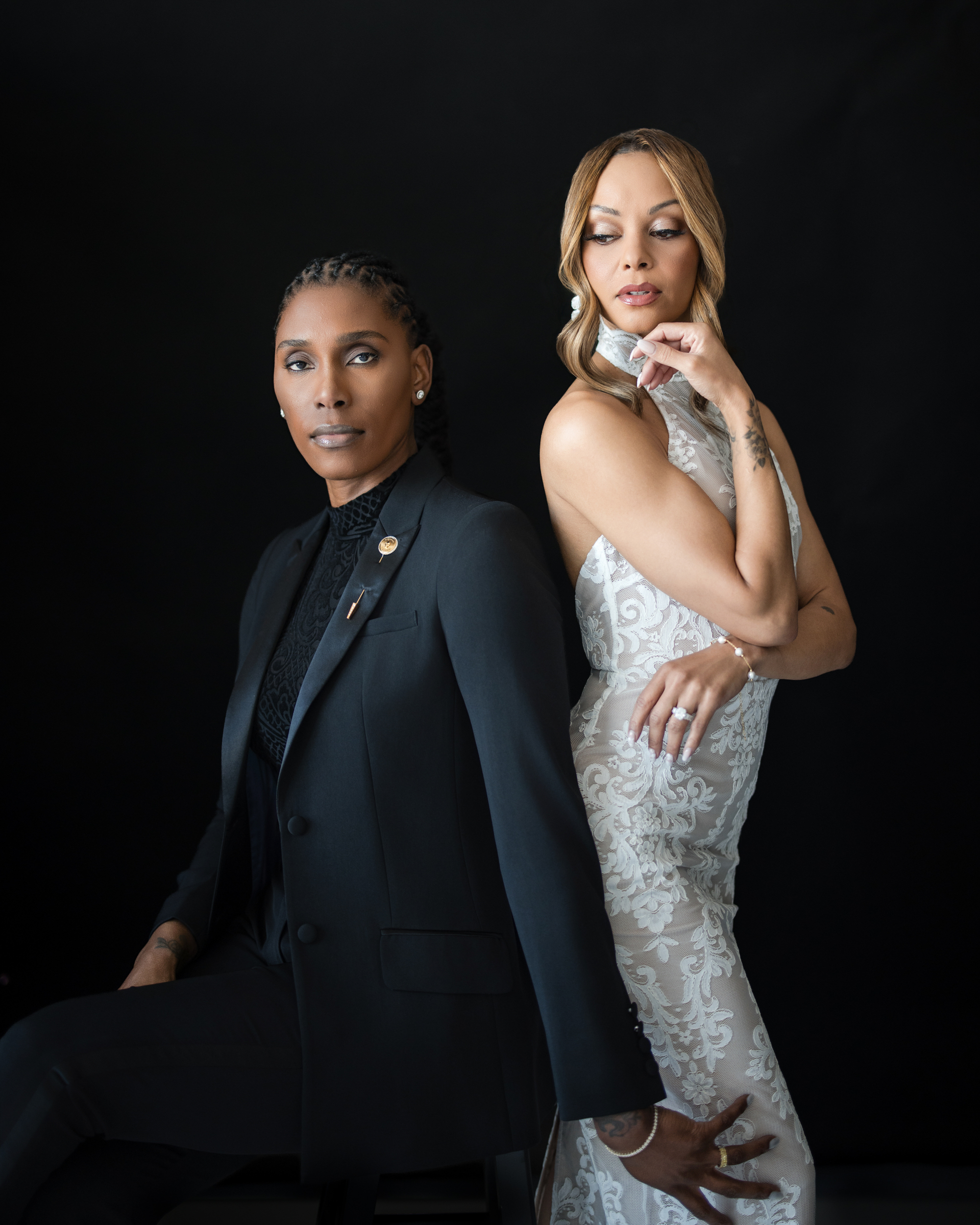 Two women pose against a black background in this striking NYC studio engagement session; one sits confidently in a black suit, while the other stands gracefully in a white lace dress, resting her hand thoughtfully on her chin.