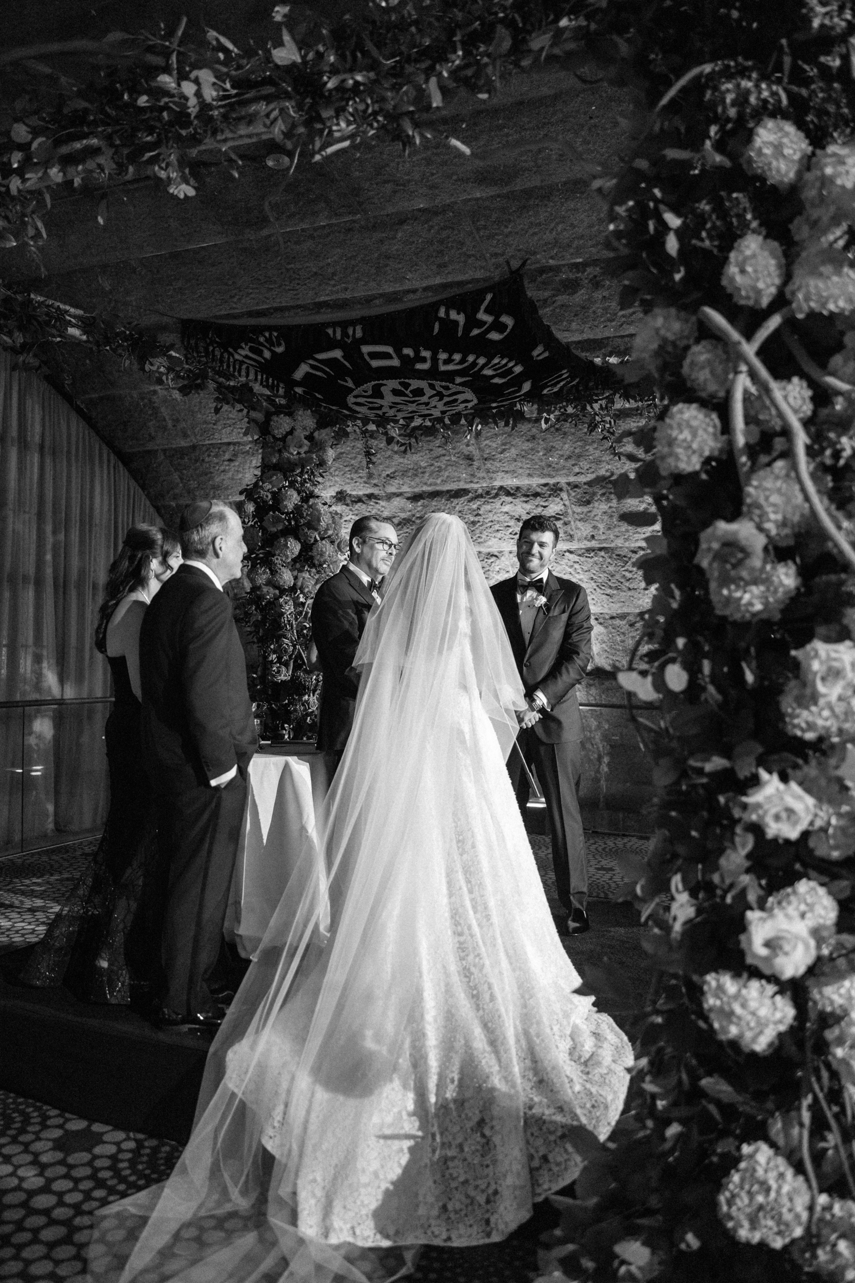 A bride and groom stand under a chuppah during a Guastavino’s wedding ceremony, surrounded by floral arrangements and a few guests, in a black-and-white photo.