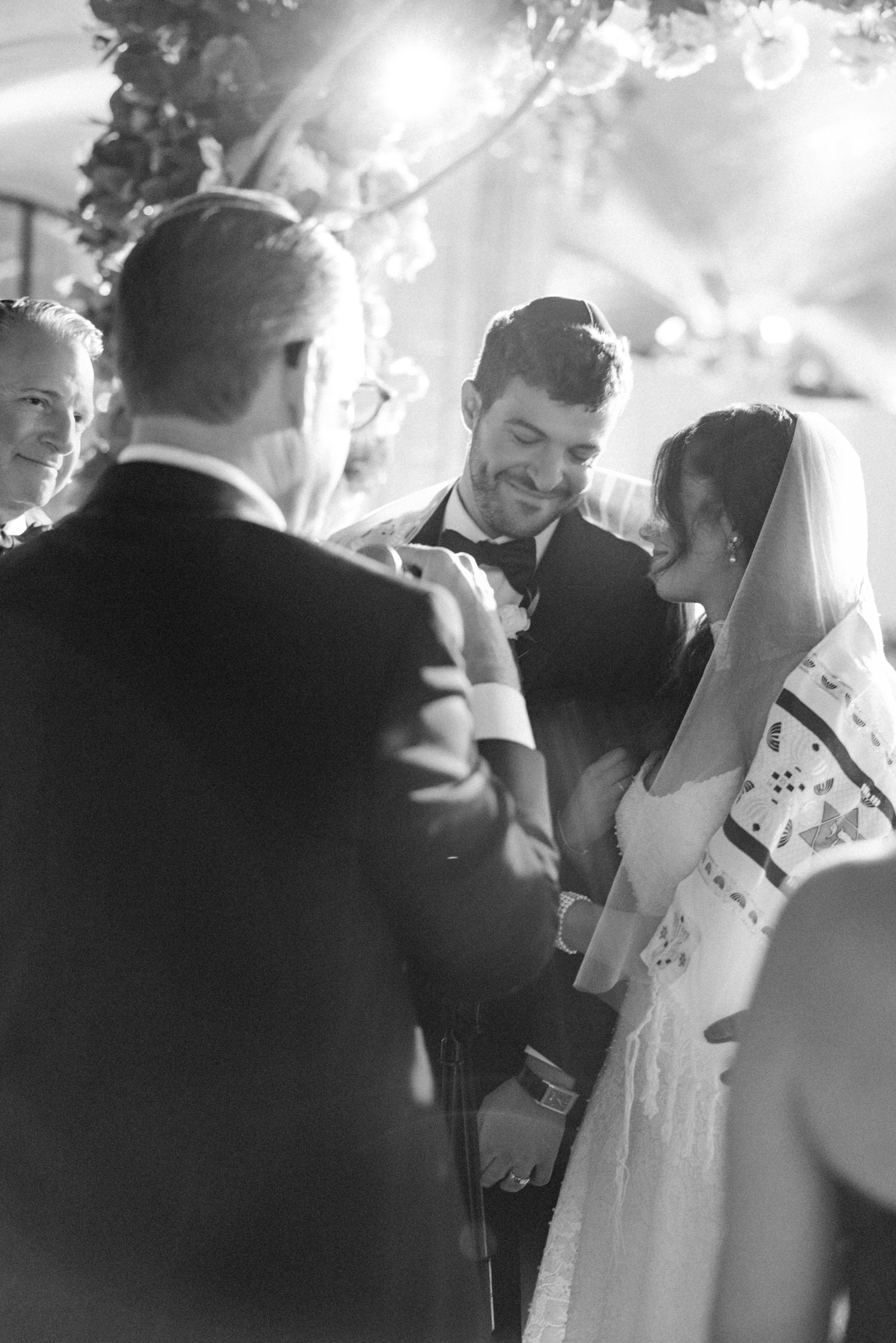 A bride and groom stand smiling under a floral canopy during a Guastavino’s wedding ceremony, surrounded by guests, in a black-and-white photo.