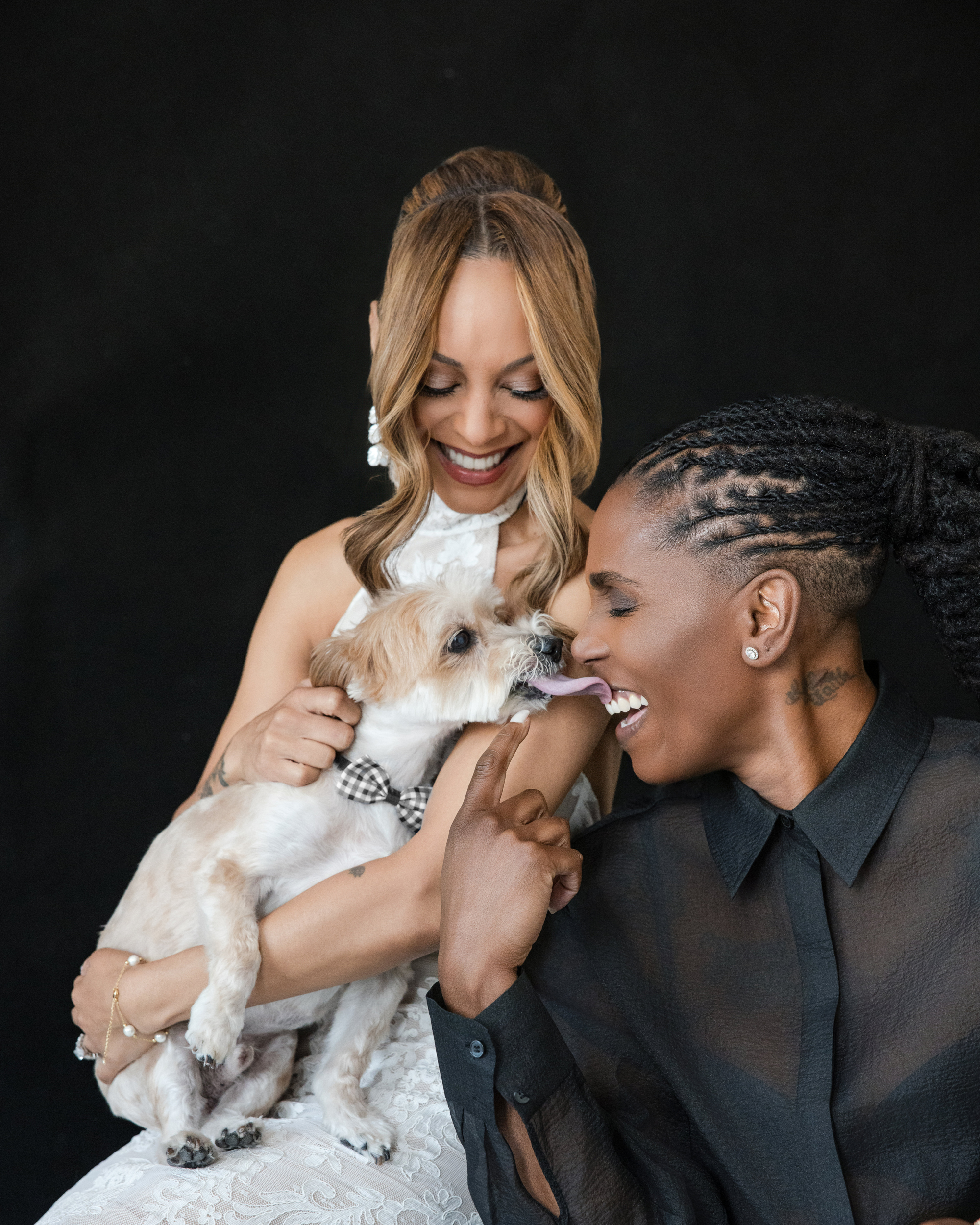 Two women smile and hold a small dog wearing a bow tie against a black background, capturing the joy of their NYC studio engagement session. One woman wears a white dress, while the other is in a sheer black shirt.