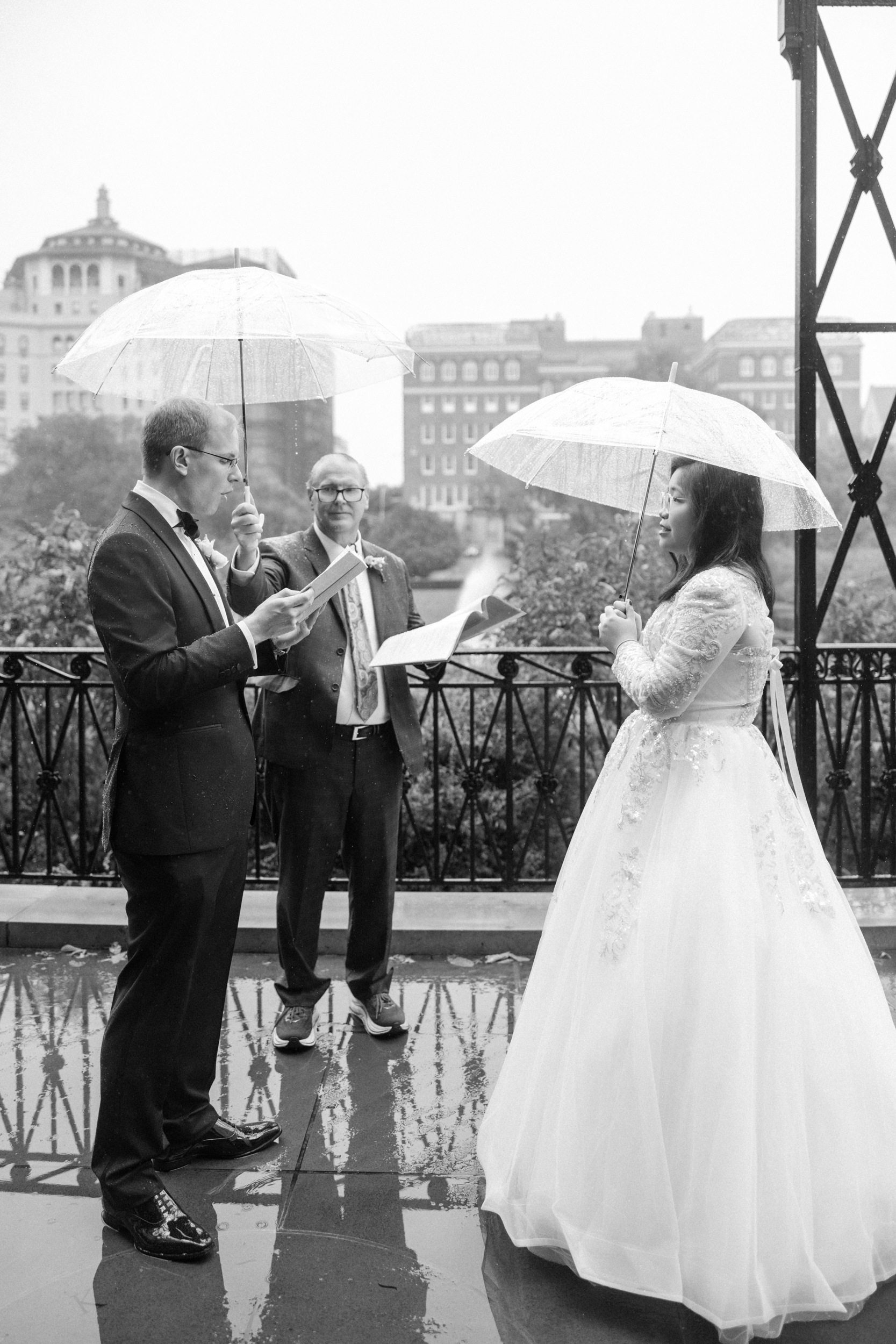 A bride and groom stand under clear umbrellas during an outdoor Conservatory Garden wedding ceremony, with an officiant between them and city buildings visible in the background.