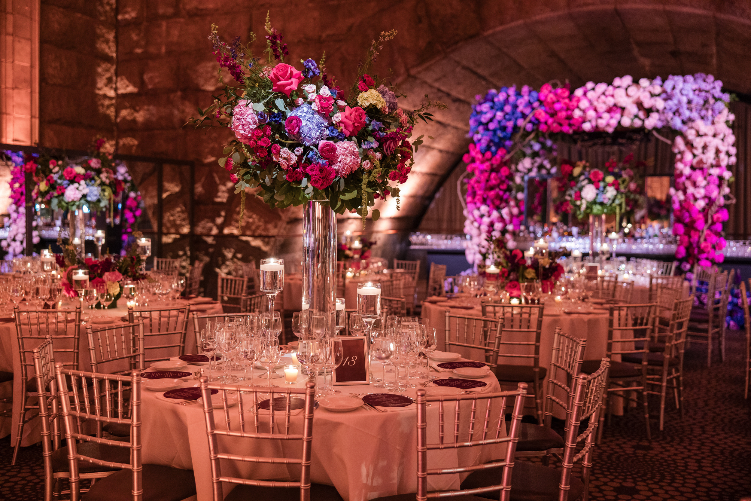 Elegant banquet hall with round tables set for an event, featuring tall floral centerpieces, glassware, and a large flower arch in the background, illuminated by warm pink and purple lighting—perfect for a sophisticated Guastavino’s wedding celebration.