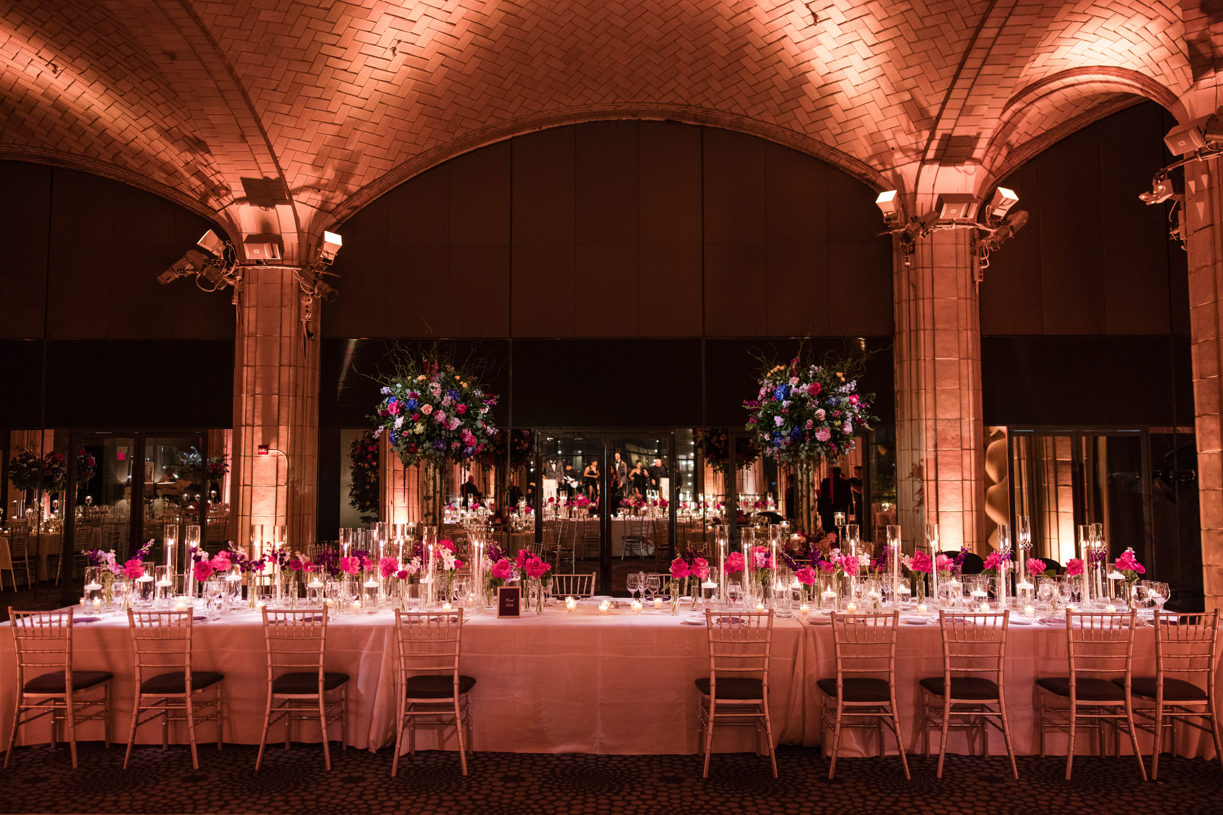 Long banquet table with white tablecloth, pink and purple flower arrangements, and candles, set in an elegant hall with arched ceilings and warm lighting—perfect for a romantic Guastavino’s wedding celebration.