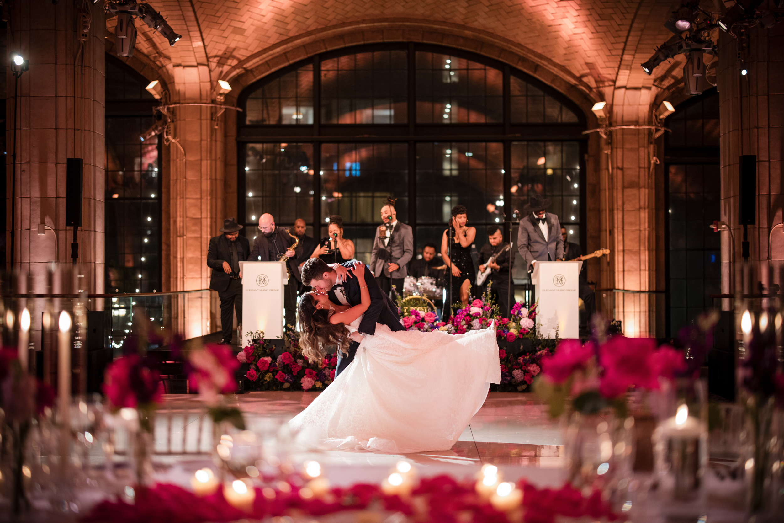 A bride and groom share a dance under warm lights in the grand hall at Guastavino’s wedding, with a live band performing on stage behind them and flower arrangements in the foreground.