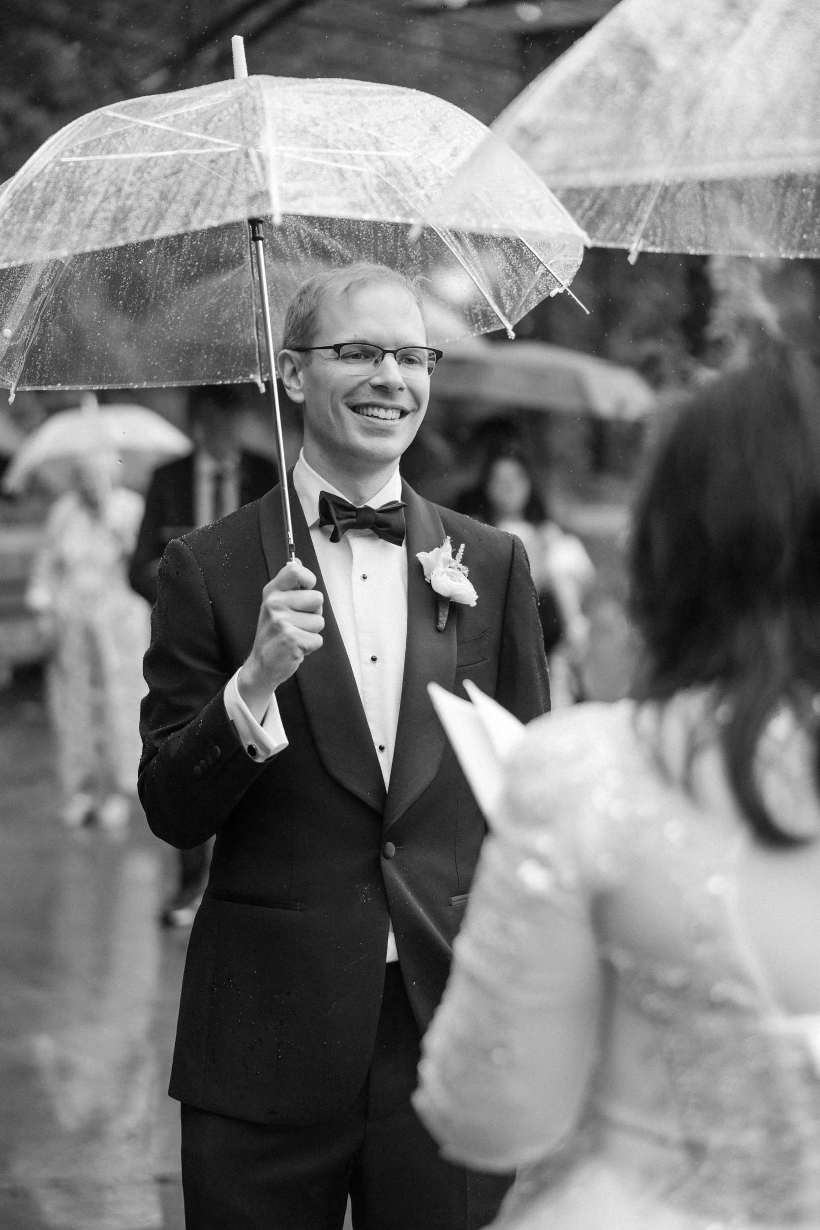 A man in a tuxedo smiles while holding a clear umbrella in the rain, facing a woman in a wedding dress during their Conservatory Garden wedding. Other people with umbrellas are visible in the blurred background.