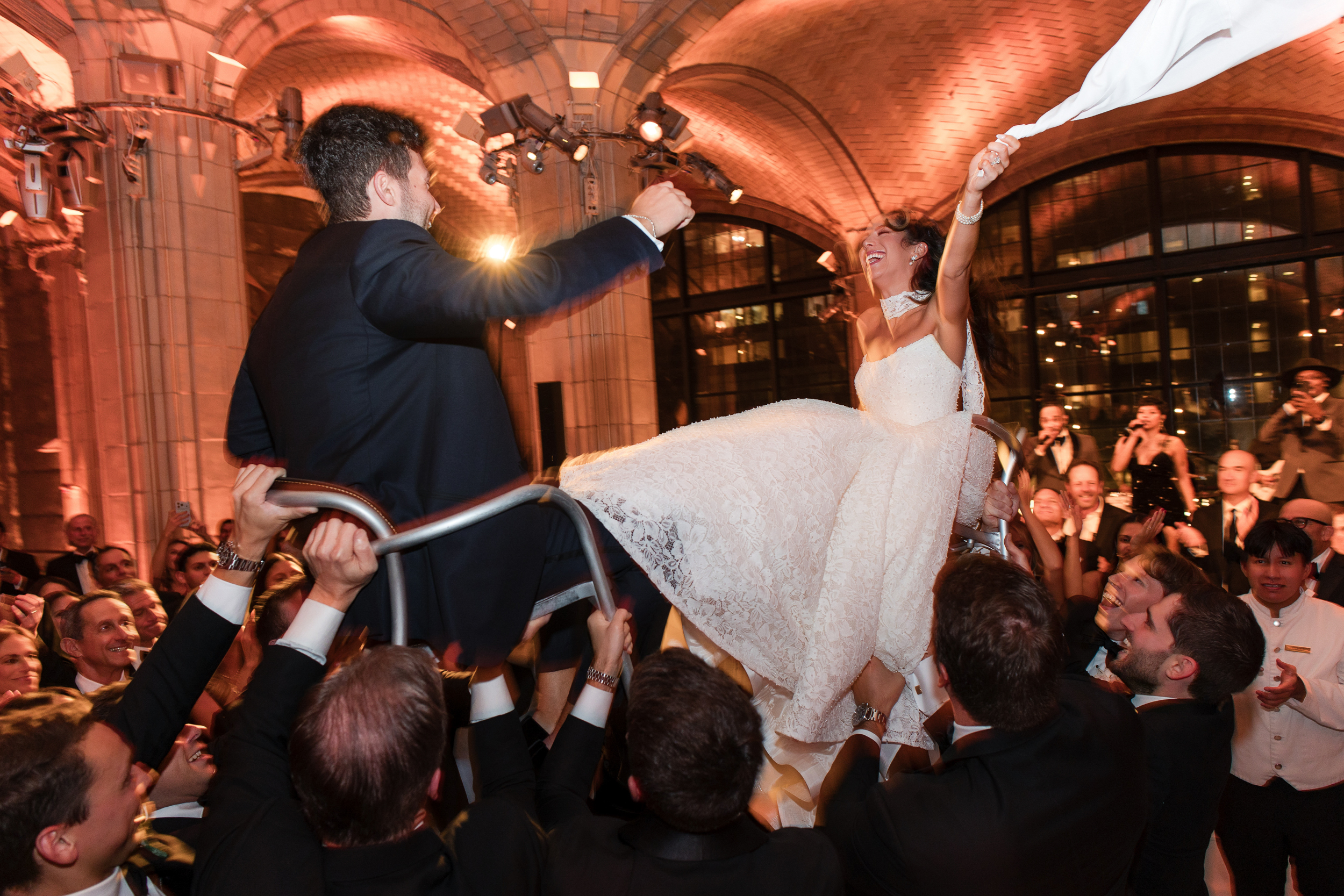 A bride and groom are lifted on chairs by guests during a lively Guastavino’s wedding celebration in a large, elegant hall.