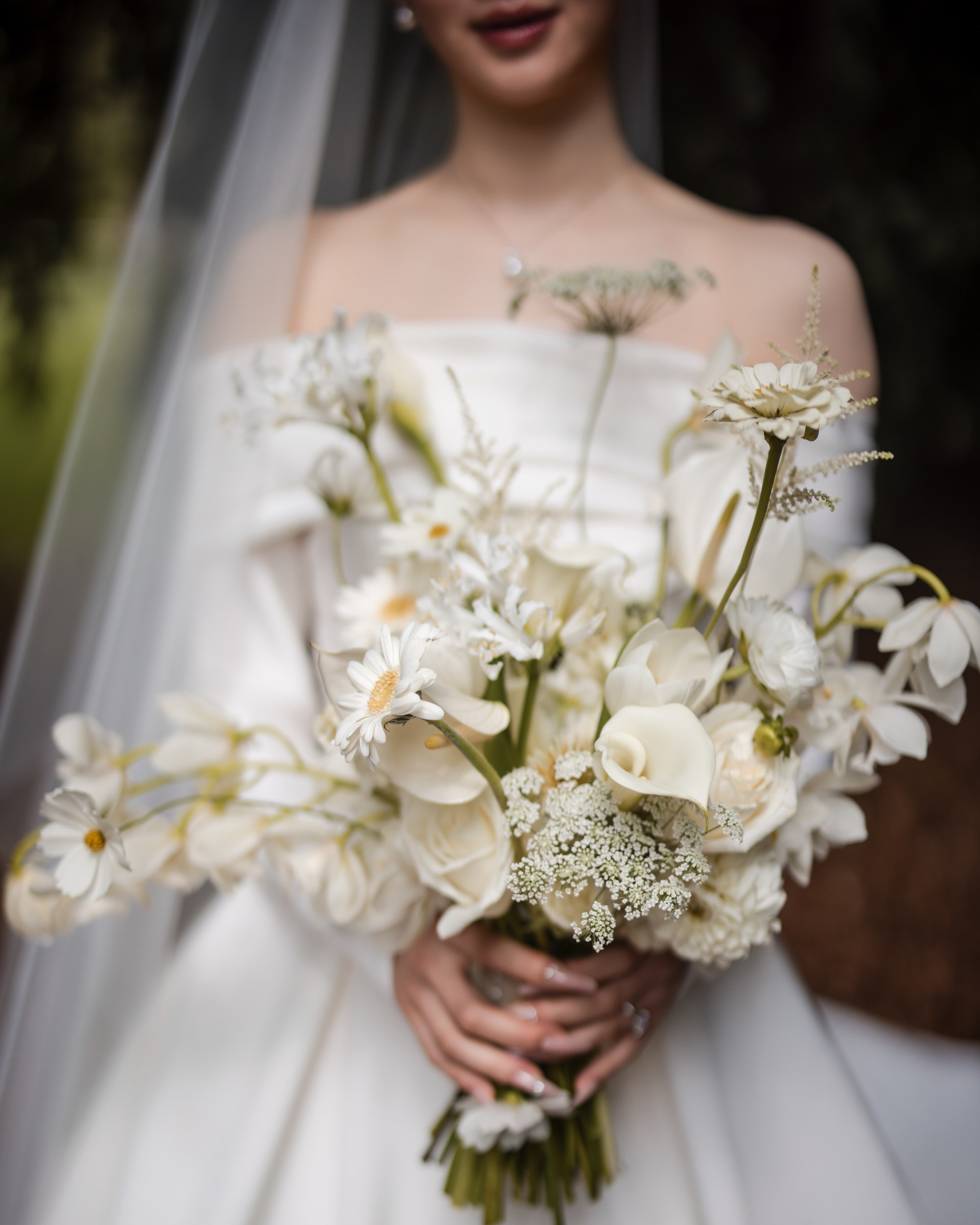 A bride in an off-shoulder white dress holds a bouquet of white flowers, including calla lilies and daisies, with a veil partially visible at her elegant Lyndhurst Mansion wedding.