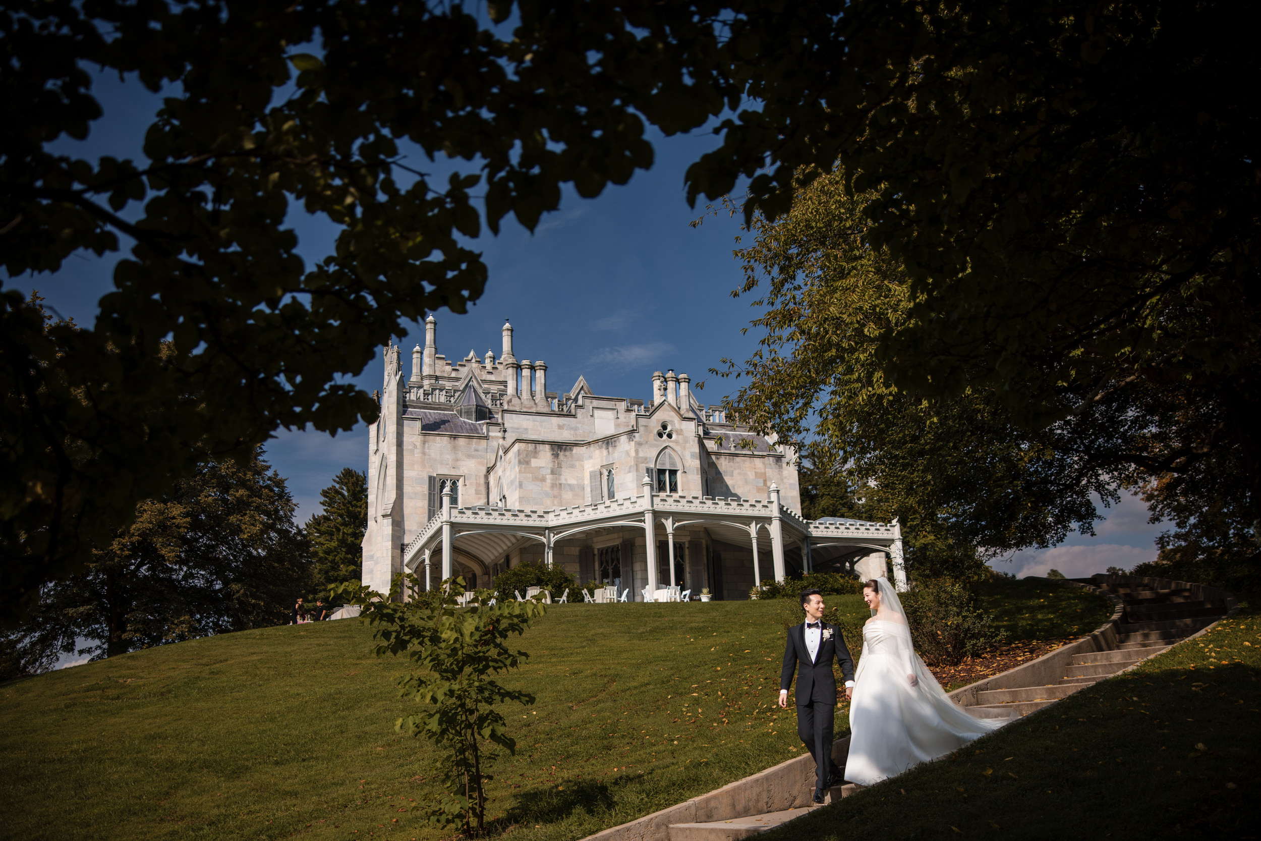 A bride and groom walk down stone steps outside the grand Lyndhurst Mansion, surrounded by trees on a sunny day, capturing the timeless elegance of a Lyndhurst Mansion wedding.