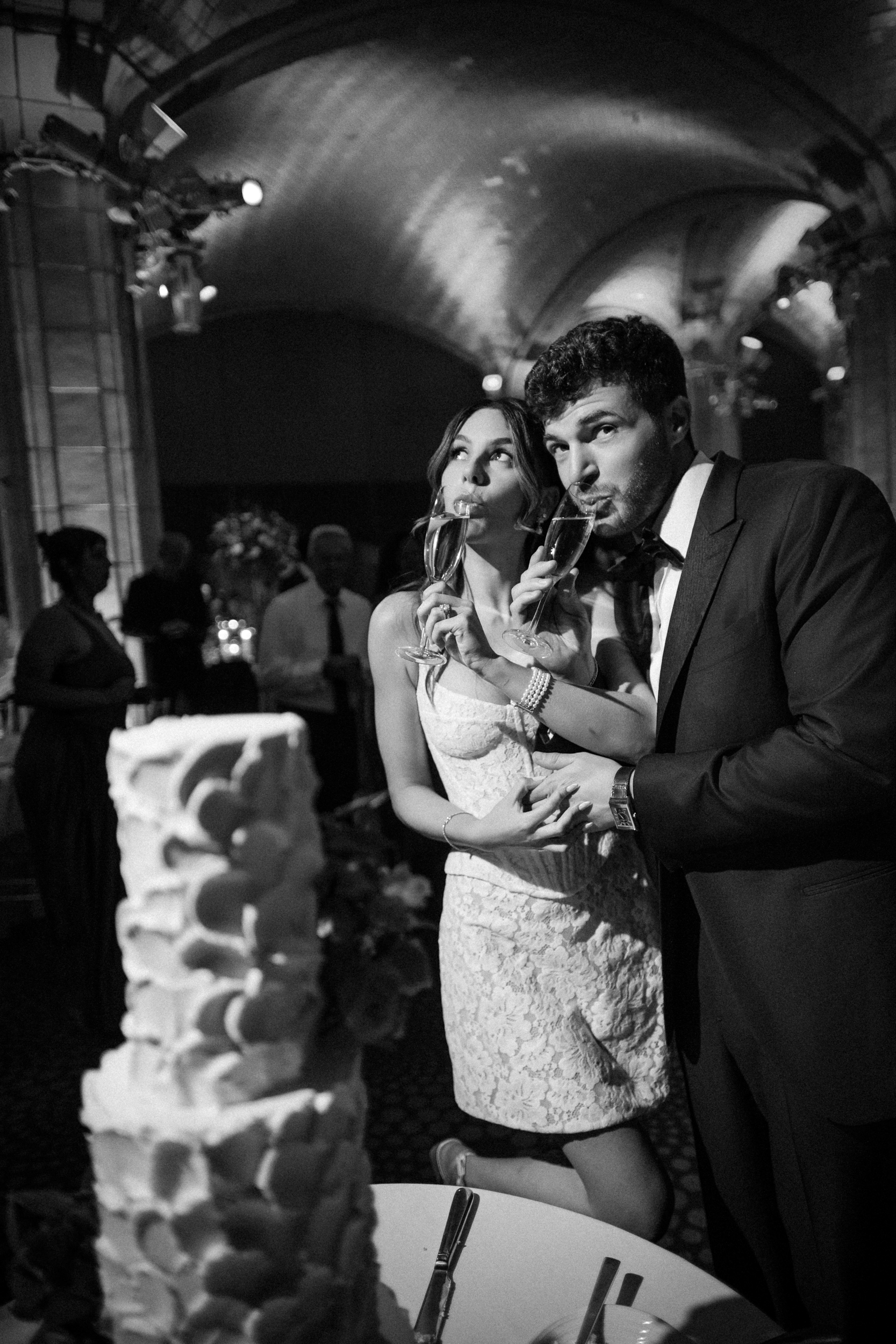 A couple in formal attire stands together eating cake with forks at a Guastavino’s wedding reception, with a tiered cake visible in the foreground.