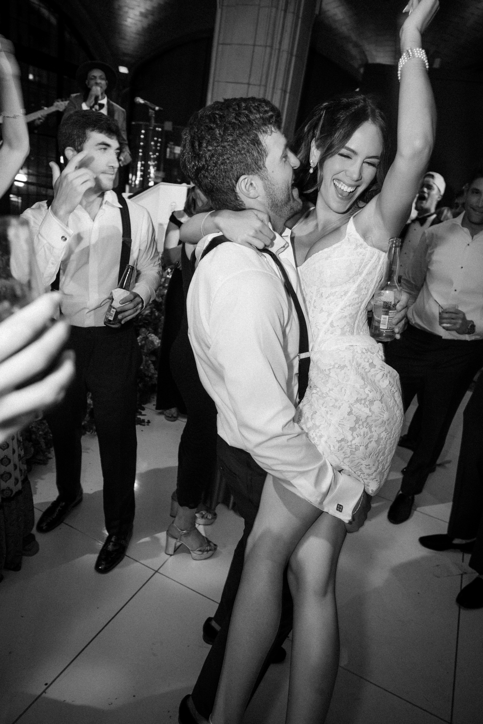 A man lifts a woman in a white dress as they smile and celebrate at a lively Guastavino’s wedding; others dance and hold drinks nearby.