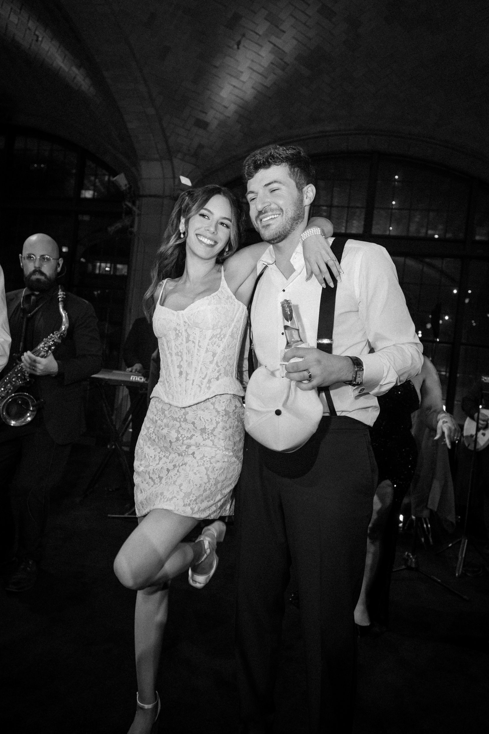 Two people pose together at a party; one holds a drink and a hat while the other rests an arm on their shoulder. A musician plays saxophone in the background, adding to the festive atmosphere of the Guastavino’s wedding. The scene is indoors at night.