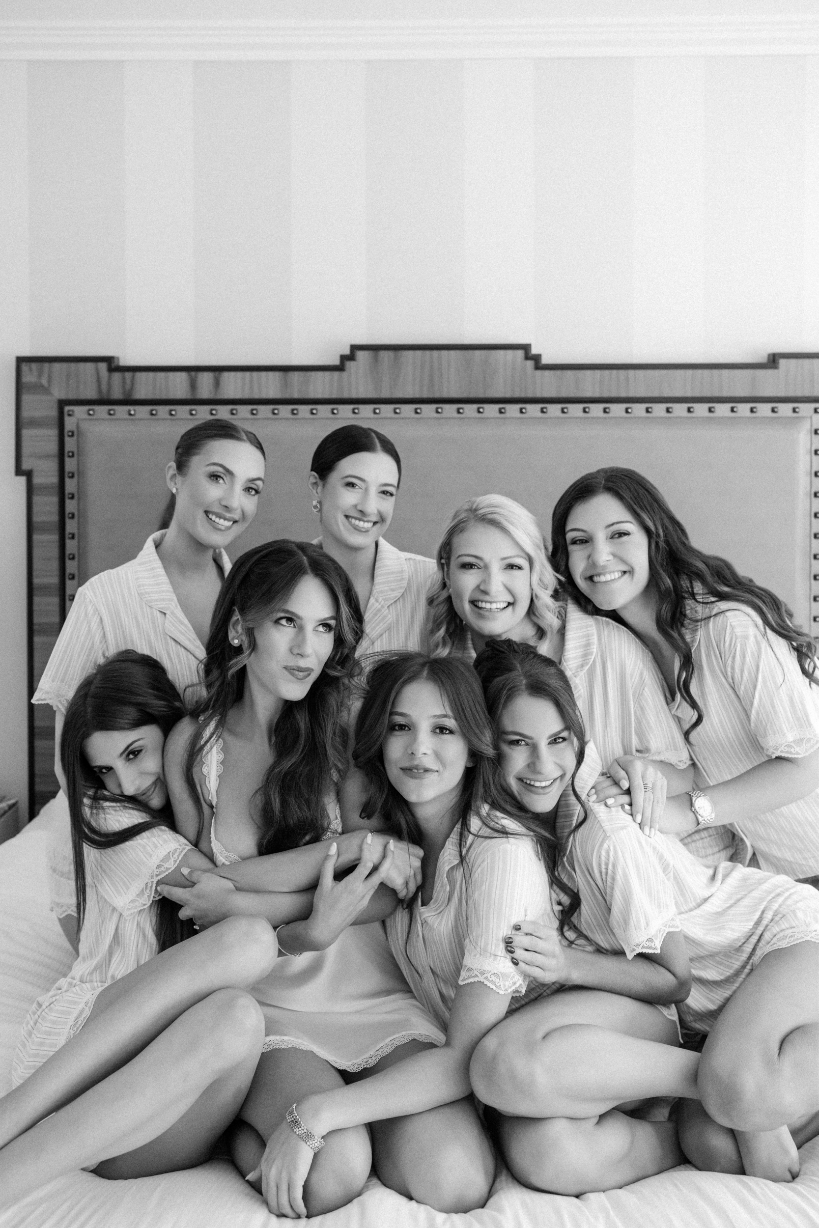 Eight women in matching pajamas sit closely together on a bed, smiling and posing for a group photo in a well-lit bedroom, sharing excitement as they get ready for their friend’s Guastavino’s wedding.