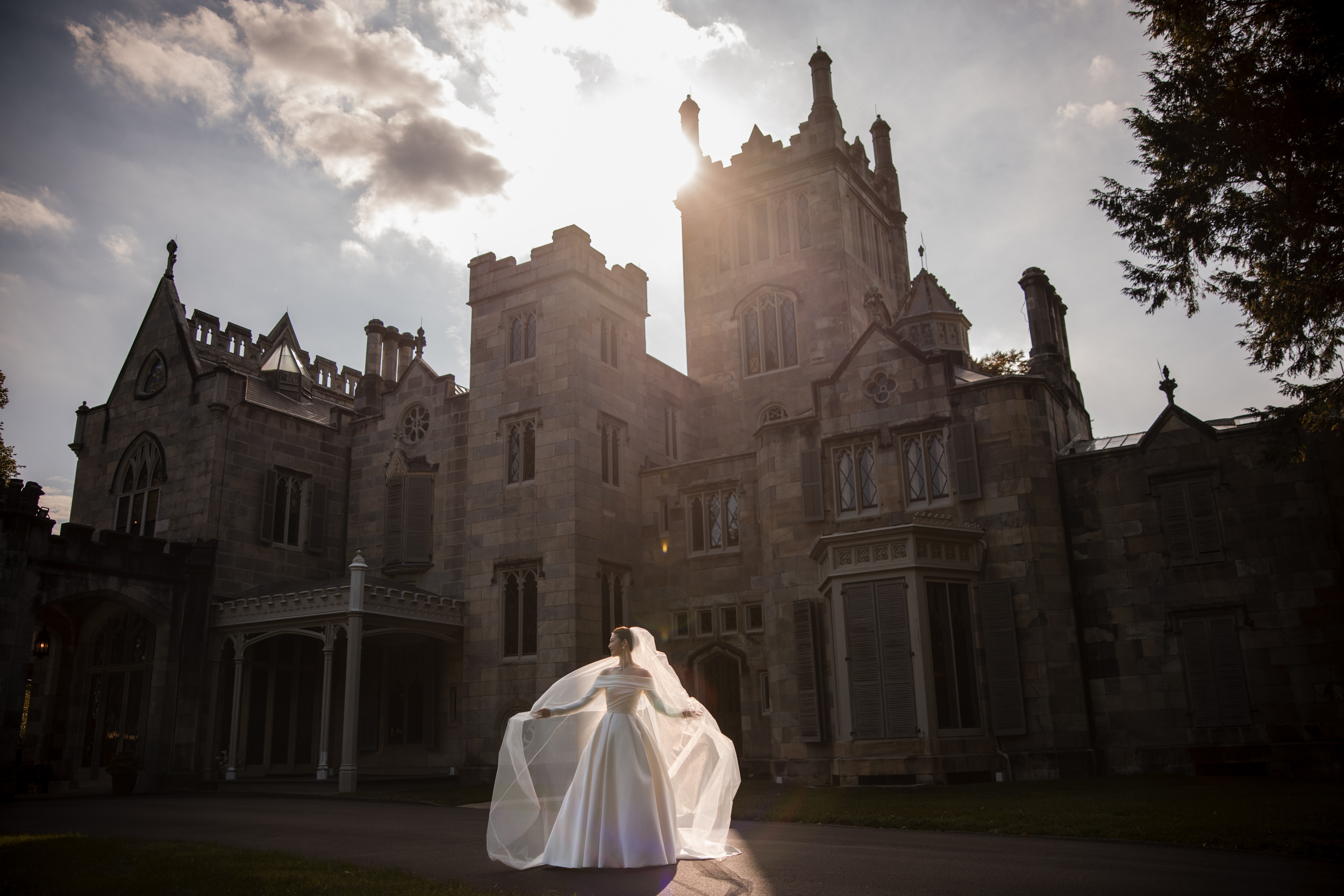 A bride in a white gown and veil stands in front of the large, historic Lyndhurst Mansion under a partly cloudy sky, with sunlight shining behind the building, capturing the elegant ambiance of a classic Lyndhurst Mansion wedding.