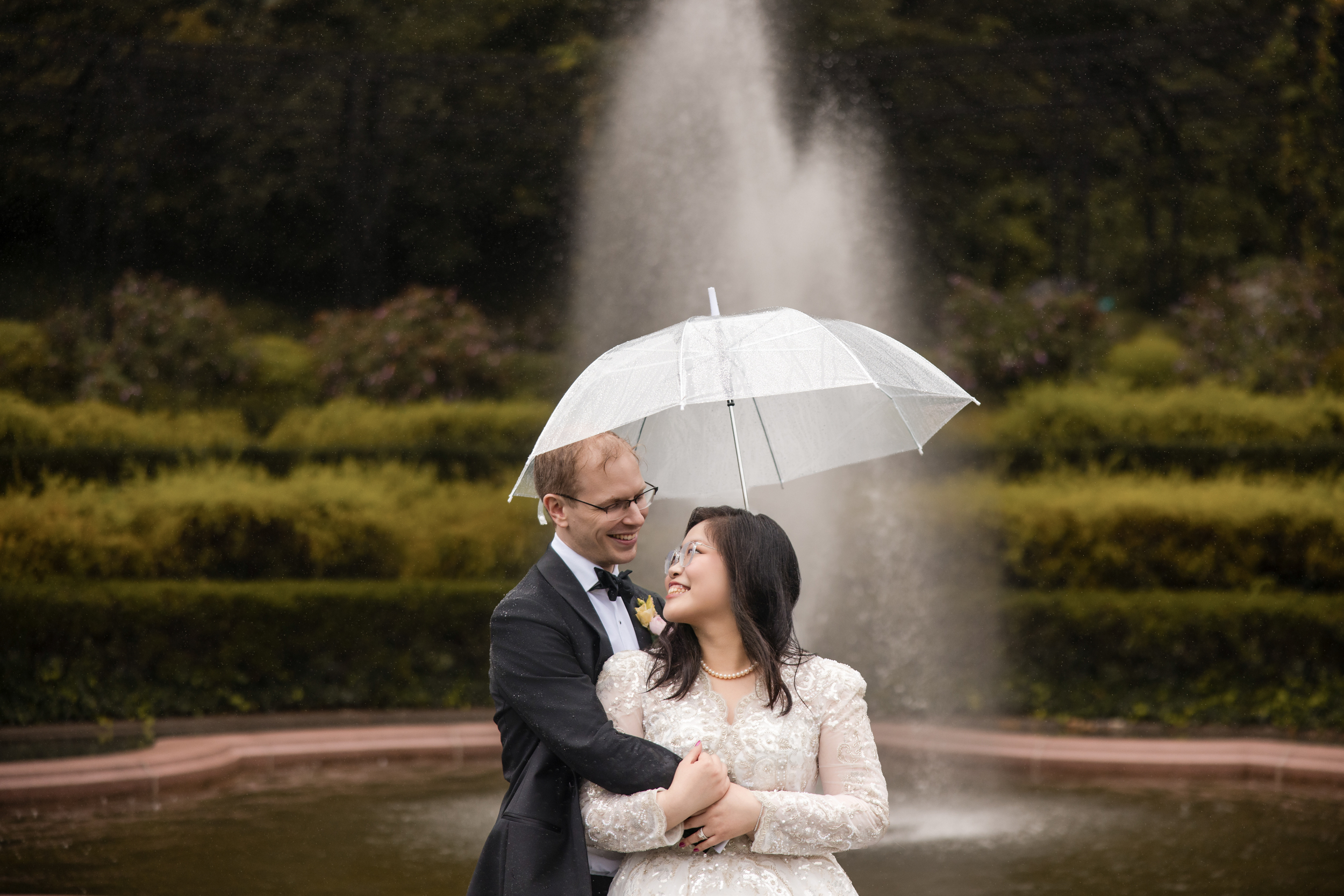 A couple in wedding attire stands in front of a fountain under a clear umbrella, smiling at each other, with green hedges and cloudy weather in the background—a timeless moment that perfectly captures the romance of a Conservatory Garden wedding.