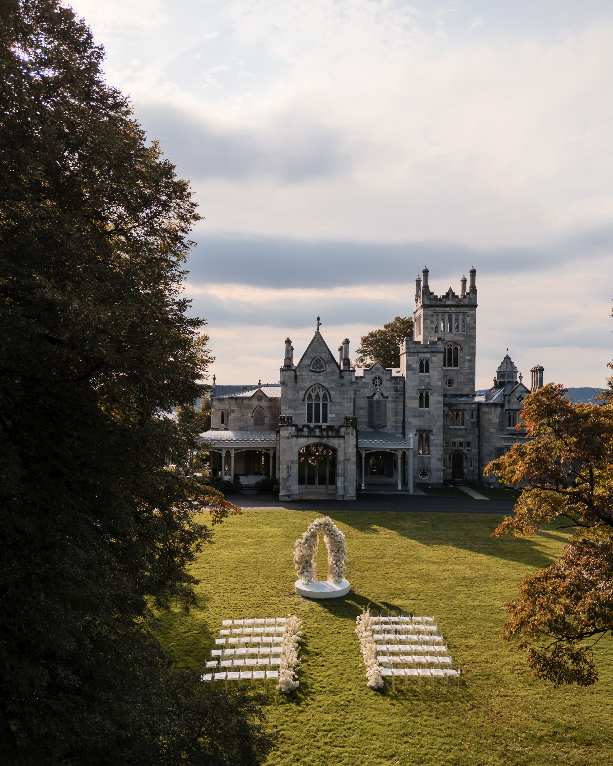 Aerial view of a Lyndhurst Mansion wedding setup with white chairs and a floral arch on a lawn in front of the large, castle-like stone building.