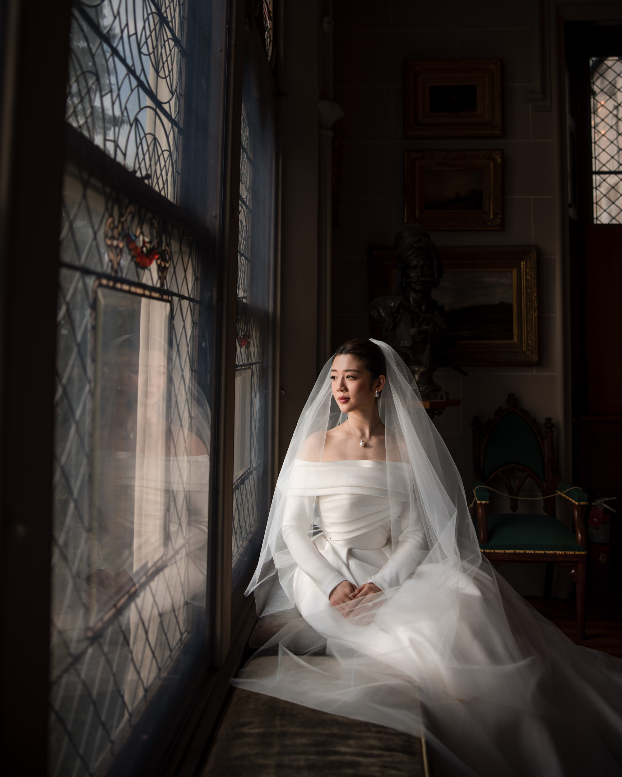 A bride in an off-the-shoulder white gown and veil sits by a window with stained glass, looking outside, in a dimly lit room with framed art and antique furniture—capturing the timeless elegance of a Lyndhurst Mansion wedding.