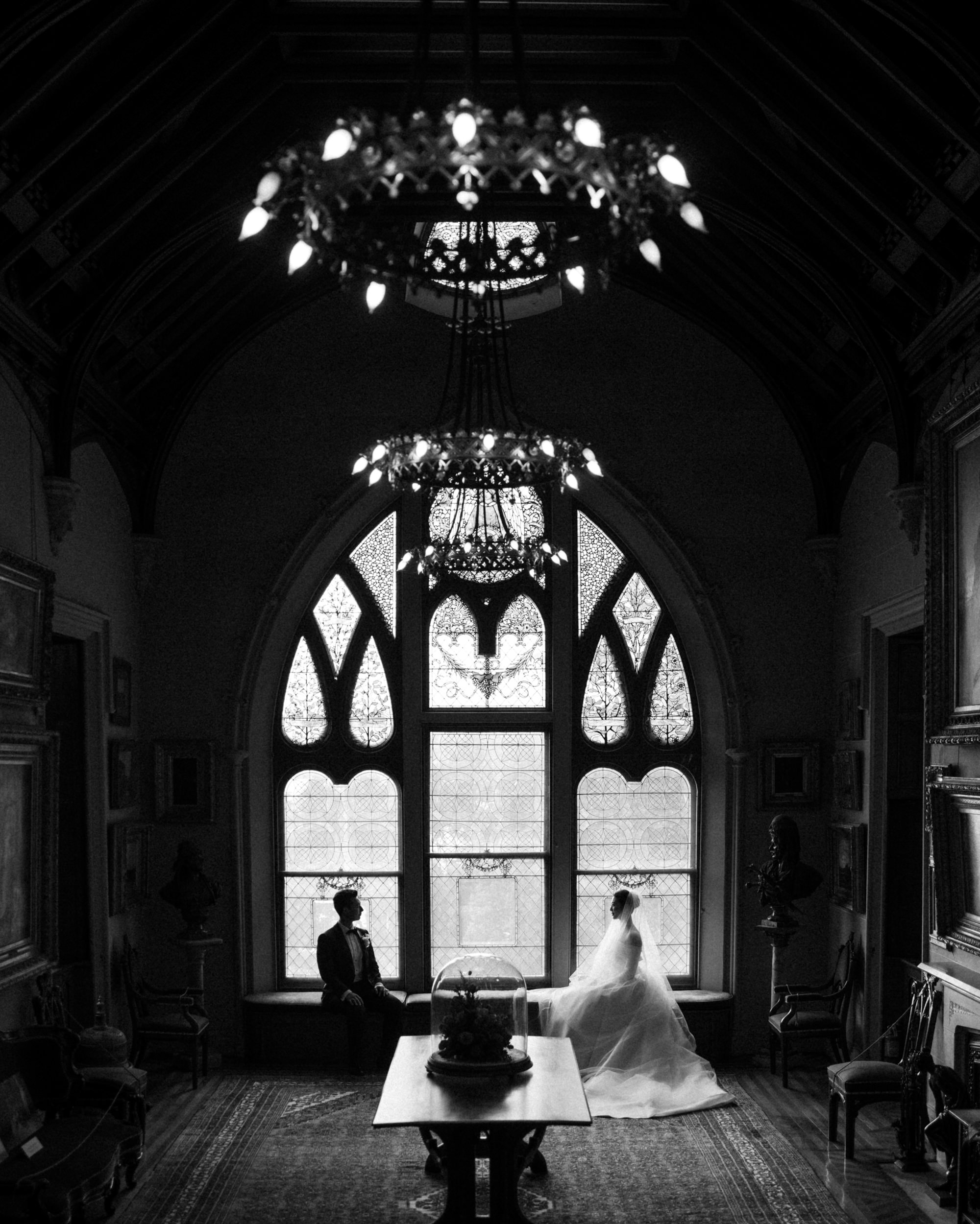 A bride and groom sit facing each other by a large arched window in a gothic-style room at their Lyndhurst Mansion wedding, illuminated by ornate chandeliers.