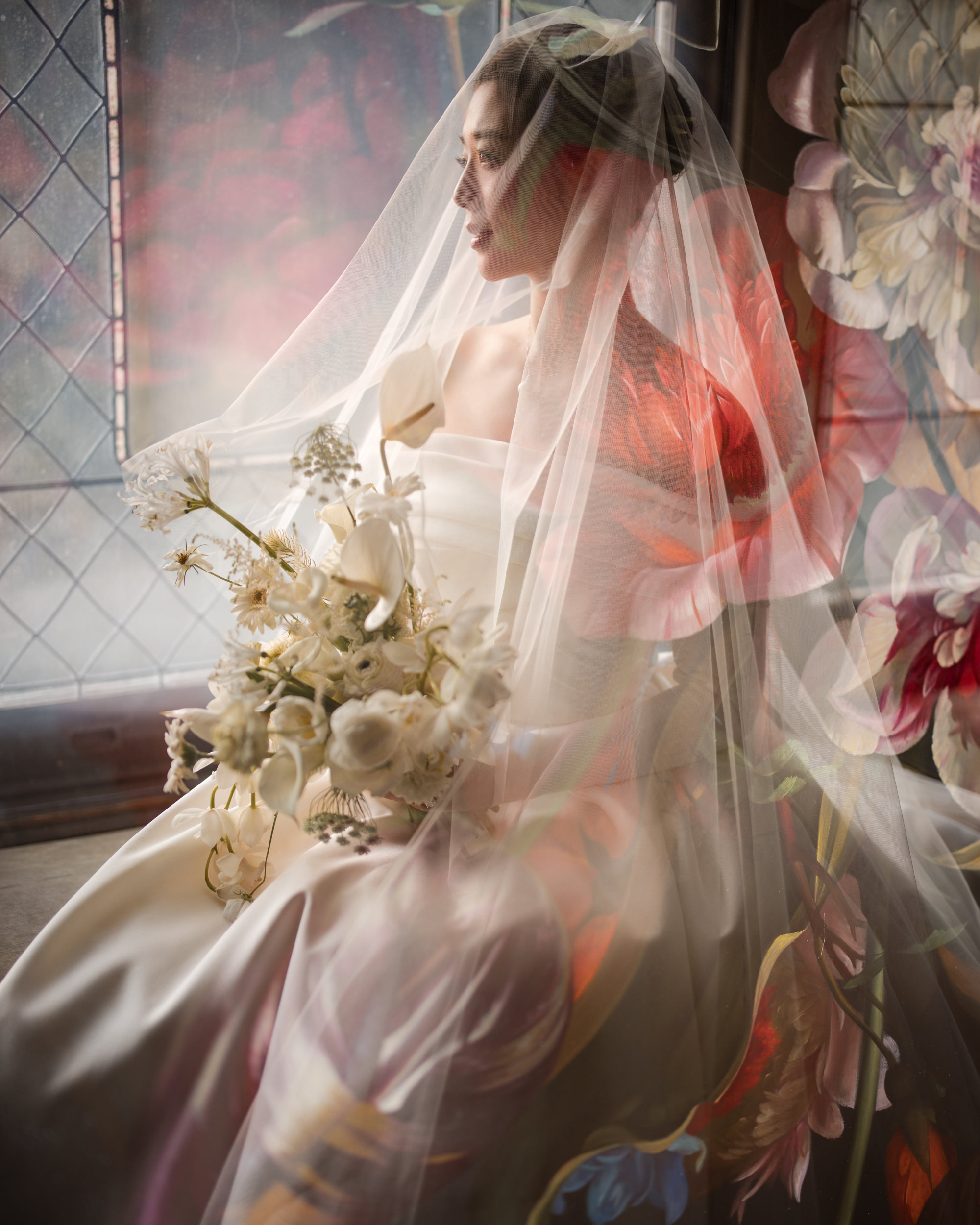 A bride in a white dress and veil holds a bouquet of white flowers, seated by a window with floral patterns and soft, artistic lighting at her romantic Lyndhurst Mansion wedding.