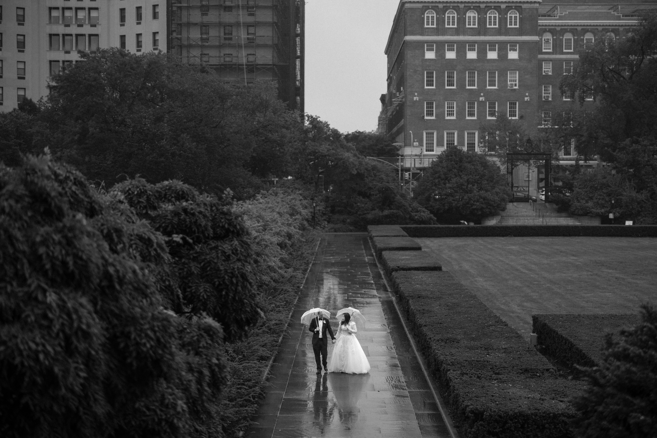 A couple in wedding attire walks under umbrellas on a wet path through Conservatory Garden, with tall buildings and dense greenery in the background.