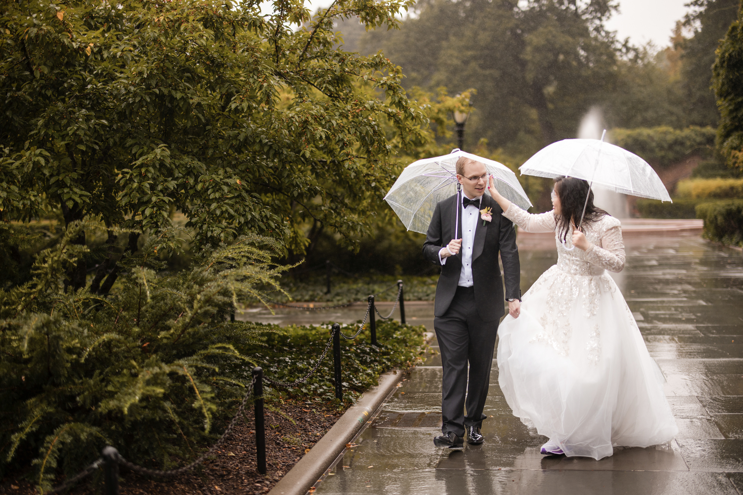 A bride and groom walk together under clear umbrellas on a rainy day in the Conservatory Garden, dressed in wedding attire.