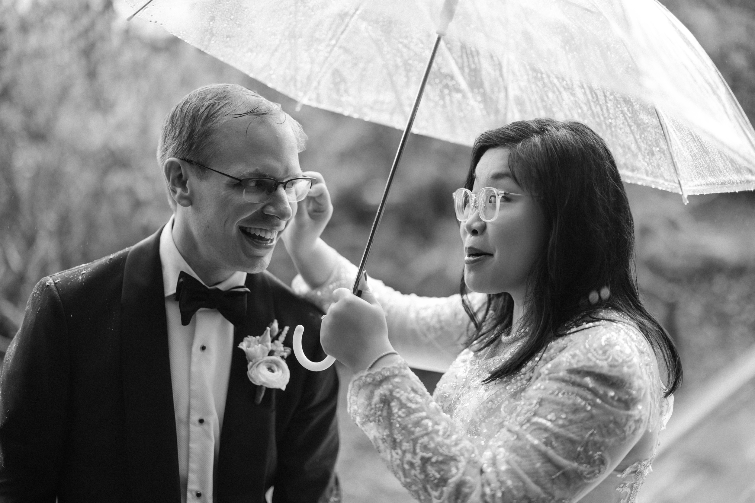 A couple dressed formally stands under a clear umbrella in the rain, sharing a joyful moment at their Conservatory Garden wedding. The woman adjusts the man’s glasses while they both smile.