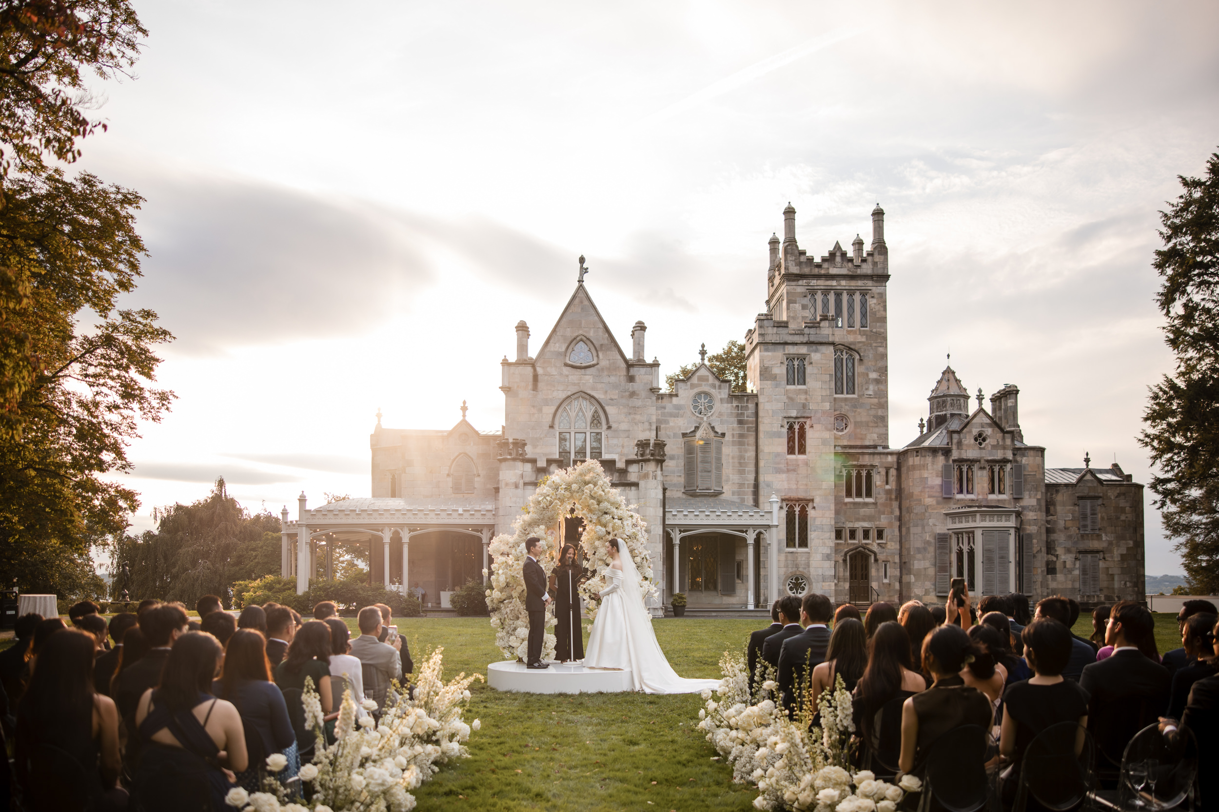 A wedding ceremony takes place outdoors in front of the grand Lyndhurst Mansion, with guests seated on either side and a floral arch elegantly framing the bride and groom.