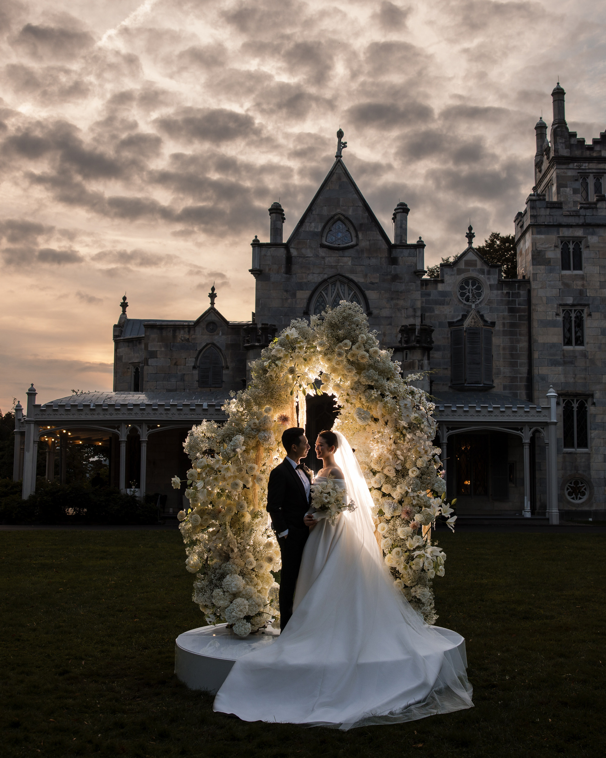 A bride and groom stand under a floral arch outside the gothic-style Lyndhurst Mansion at sunset, facing each other and holding a bouquet.