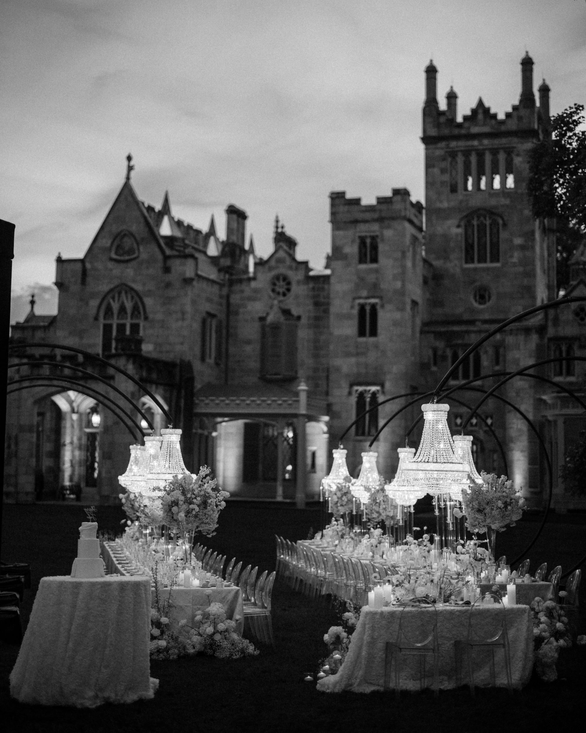 Elegant outdoor dinner setup with chandeliers and floral arrangements in front of a large, historic stone building at dusk, perfect for a romantic Lyndhurst Mansion wedding.