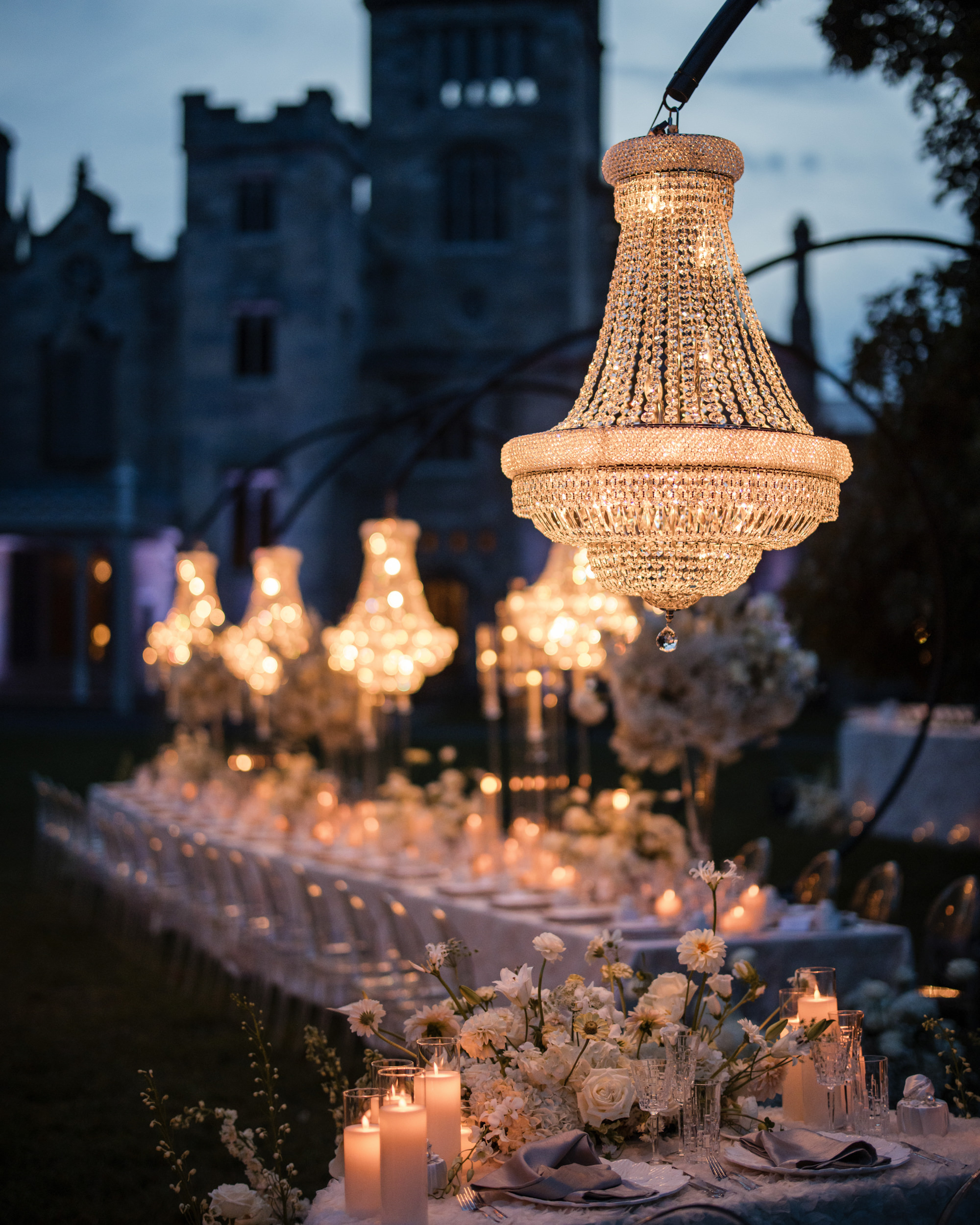 An elegant outdoor dinner setup with a long table, white flowers, candles, and chandeliers is beautifully arranged in front of a large stone building at dusk—perfect for a romantic Lyndhurst Mansion wedding.