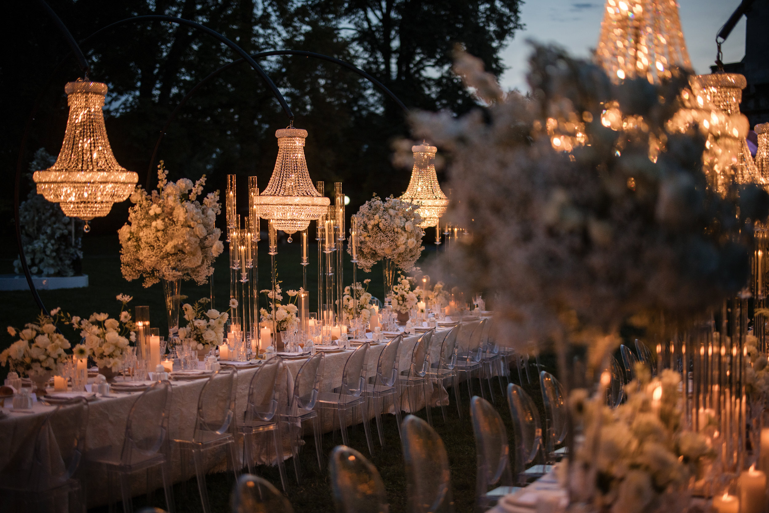 Elegant outdoor dinner setup at a Lyndhurst Mansion wedding, with long tables, white floral arrangements, clear chairs, tall candles, and chandeliers illuminated at dusk.