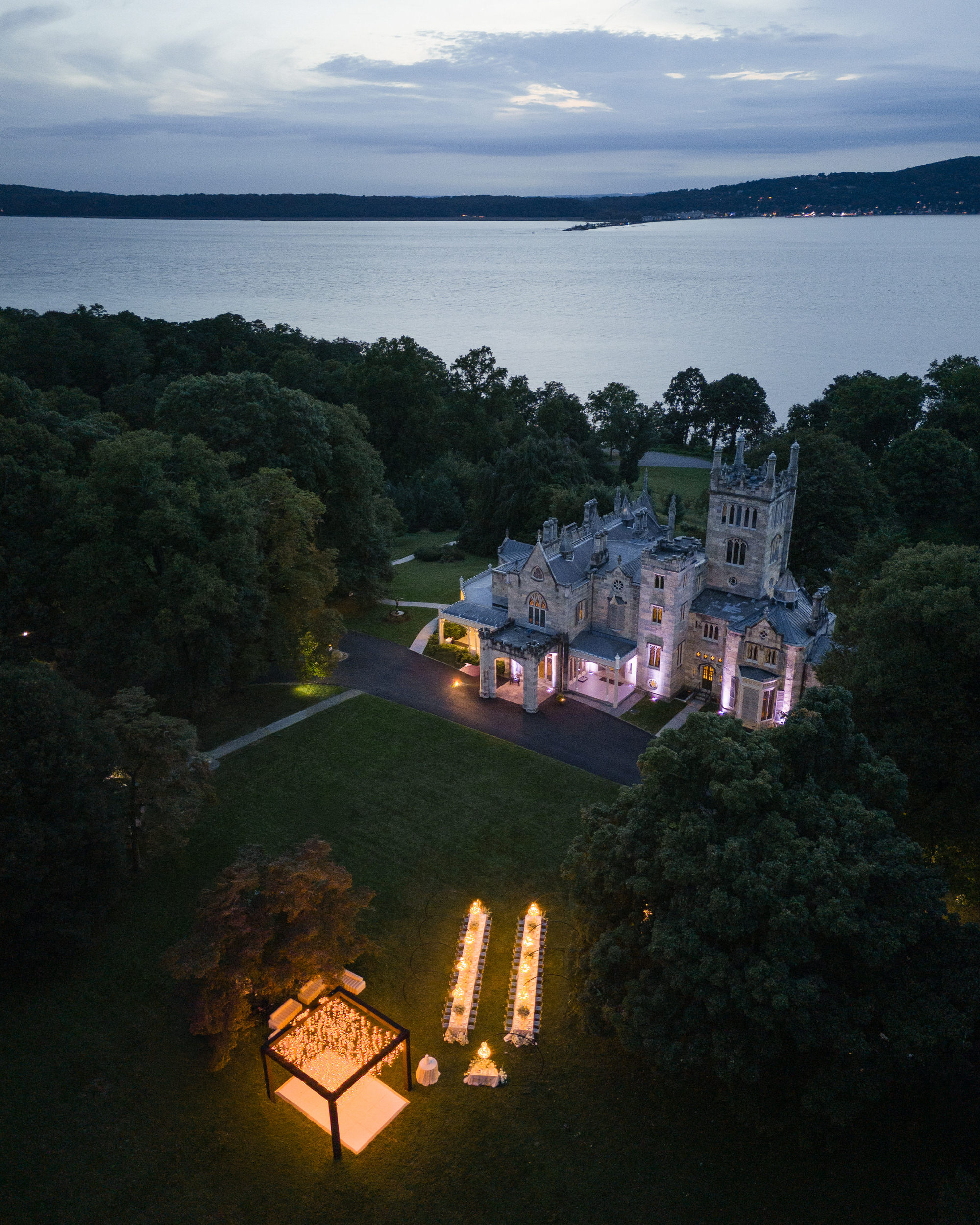 Aerial view of an illuminated castle-like building by a lake at dusk, with outdoor dining tables and a lit tent set up on the lawn, creating a romantic ambiance reminiscent of a Lyndhurst Mansion wedding.