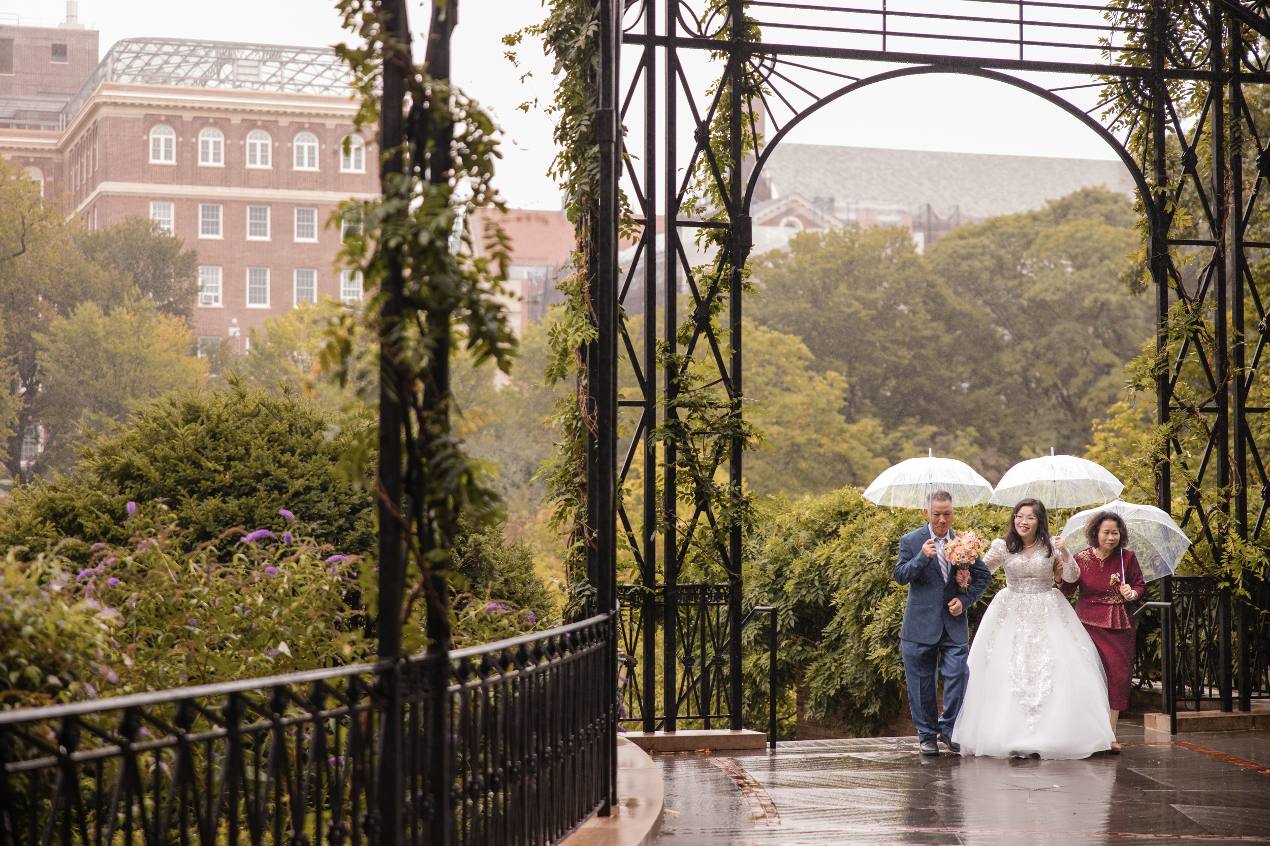 A bride walks outdoors between two people holding umbrellas on a rainy day, with greenery and a building visible in the background, capturing the elegance of a Conservatory Garden wedding.