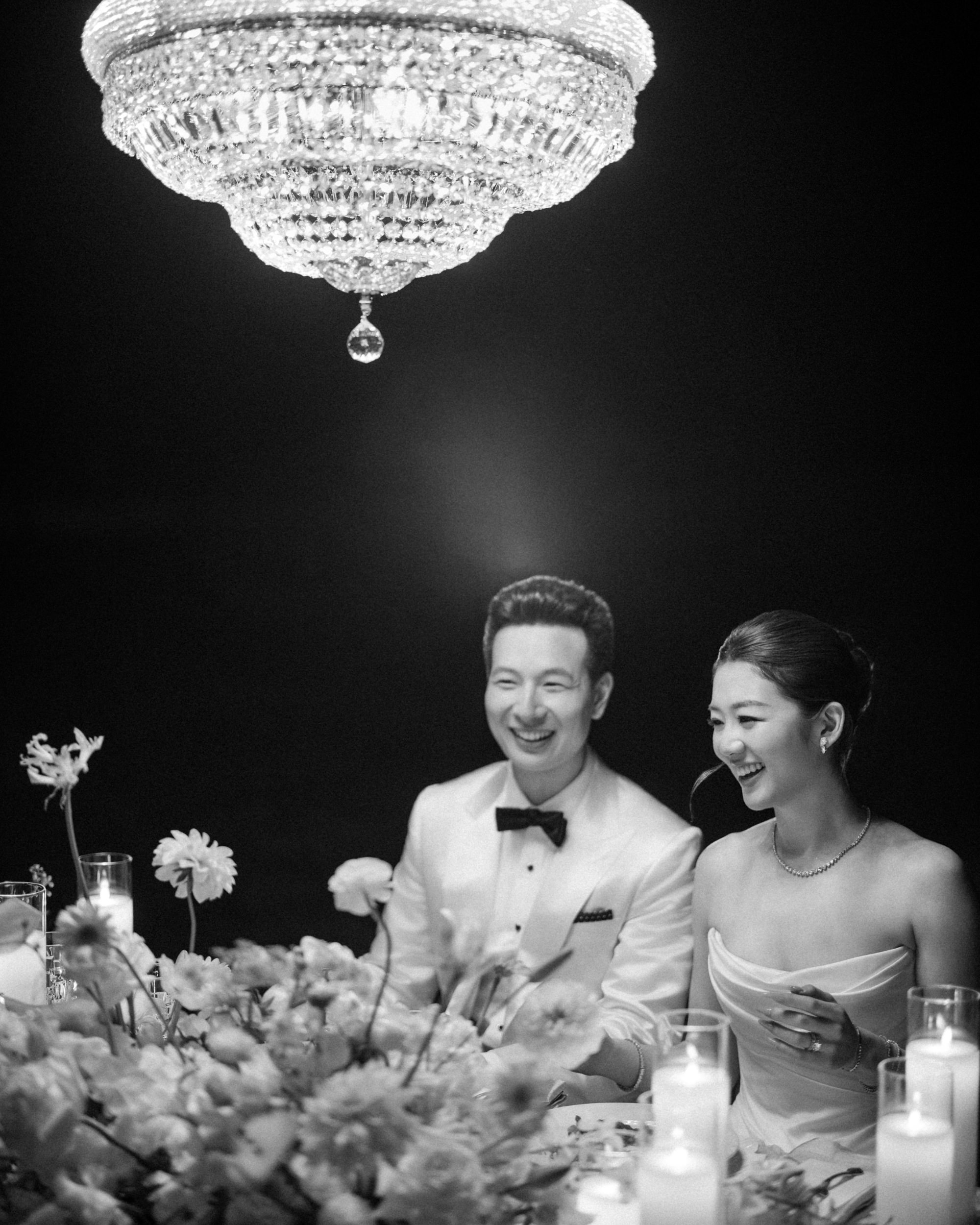 A couple dressed formally sits at a candlelit table with flowers, smiling and laughing beneath a large chandelier in a dimly lit room, capturing the timeless romance of a Lyndhurst Mansion wedding.