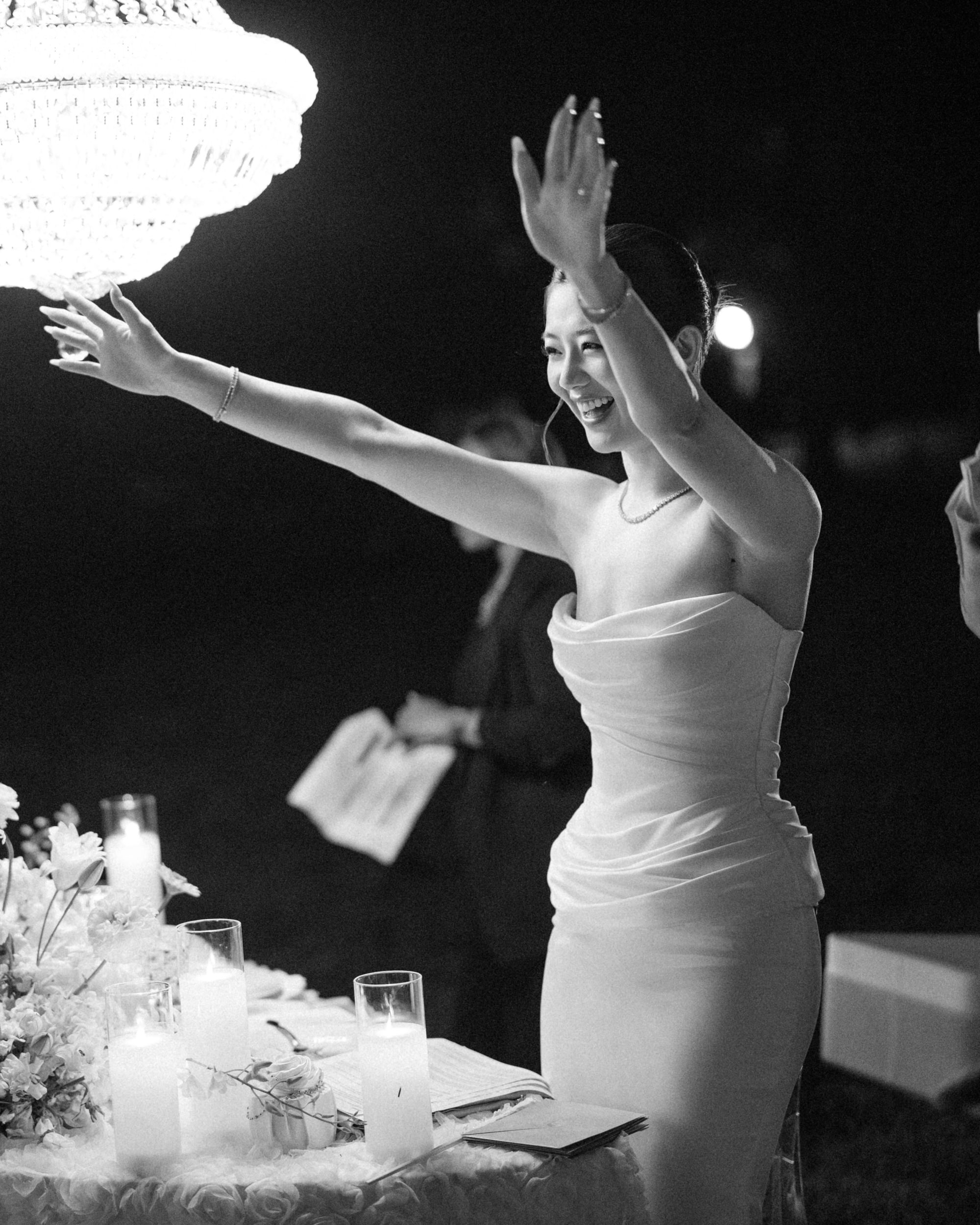 A woman in a strapless dress smiles and raises her arms near a table with candles and flowers, under a chandelier at night during a magical Lyndhurst Mansion wedding.