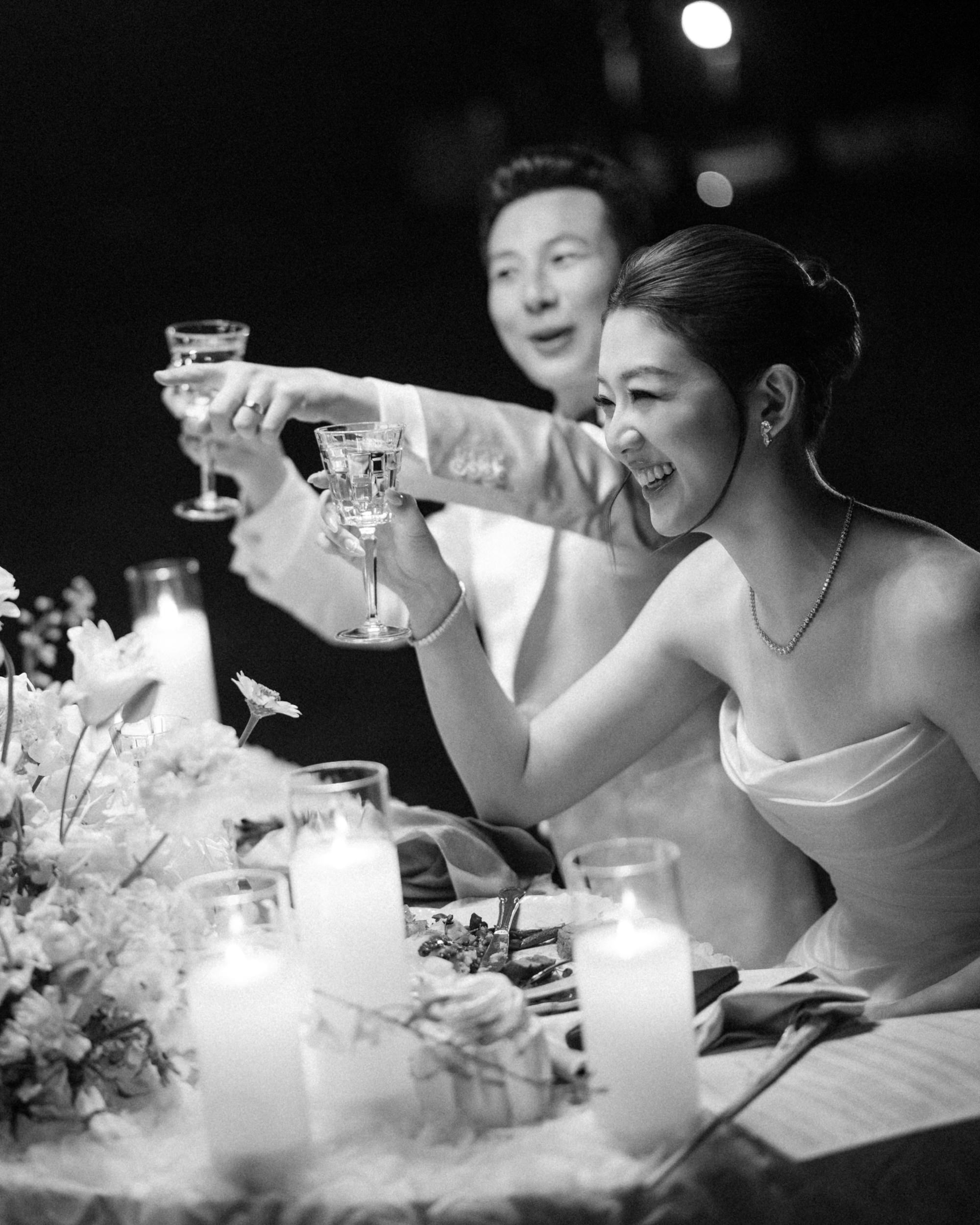 A couple in formal attire sits at a decorated table with candles and flowers, smiling and raising glasses in a toast during their elegant Lyndhurst Mansion wedding.