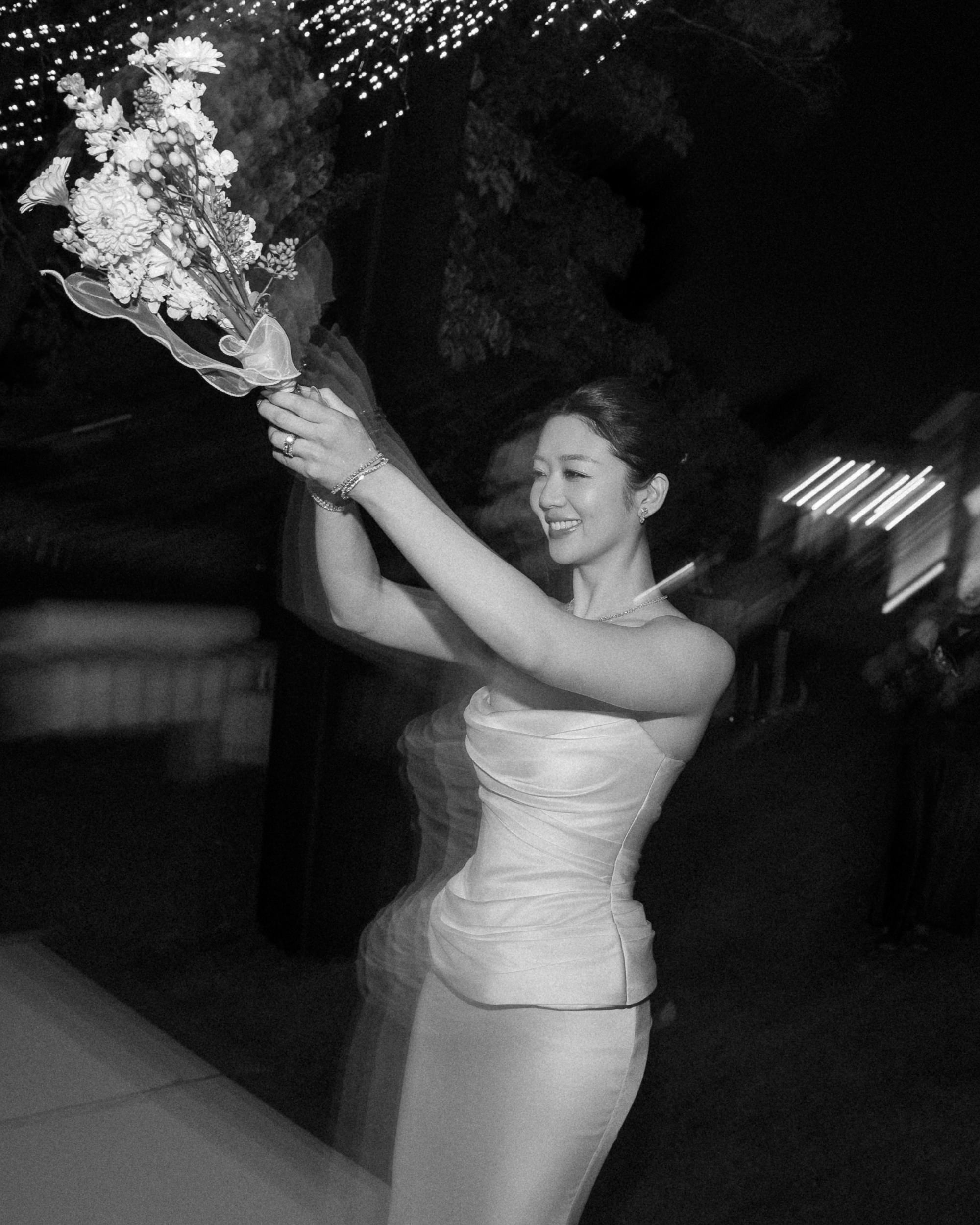 Woman in a sleeveless dress holding a bouquet of flowers, preparing to toss it at a Lyndhurst Mansion wedding, with lights and people blurred in the background.