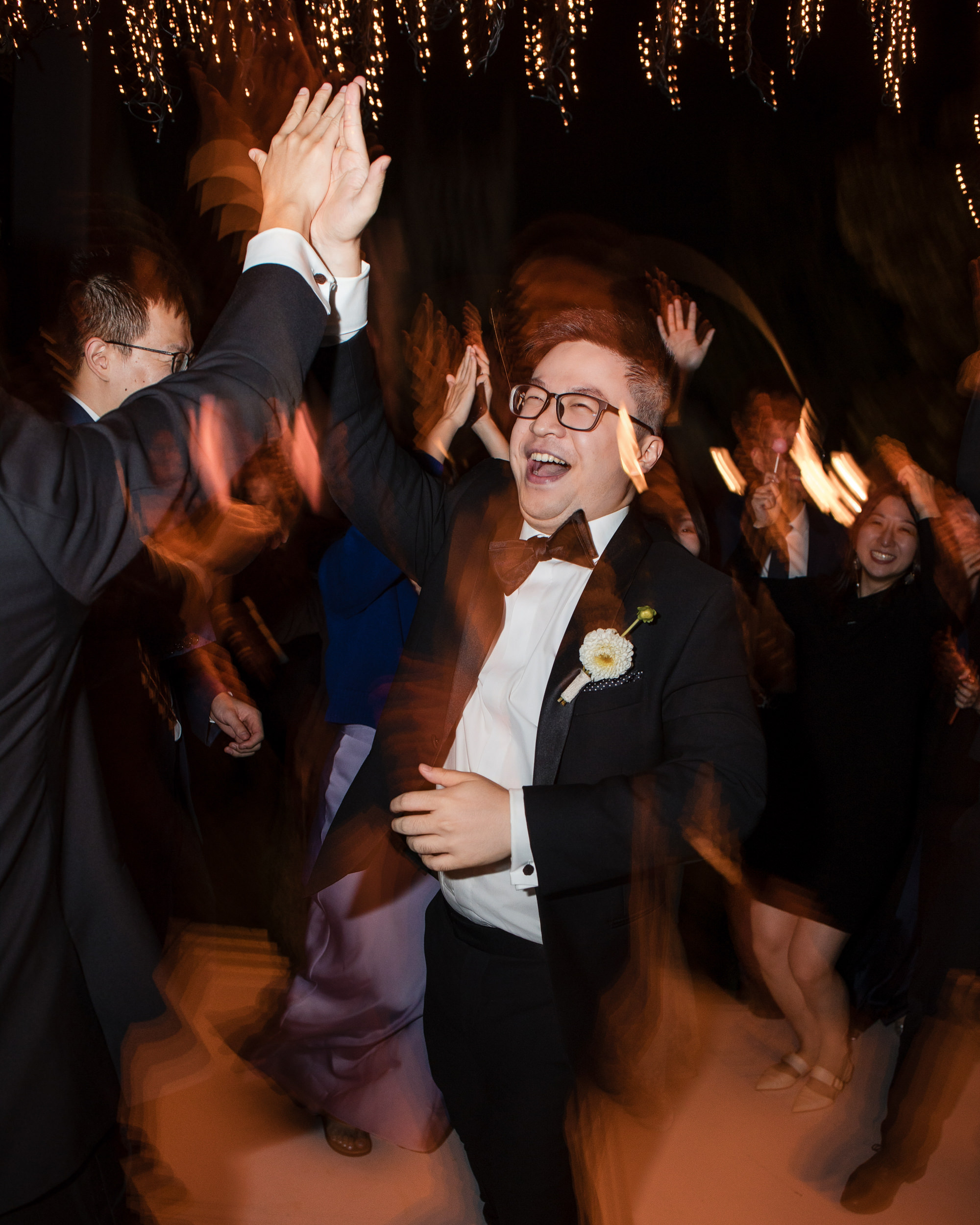A man in a tuxedo smiles and high-fives someone at a lively Lyndhurst Mansion wedding, surrounded by people and string lights, with motion blur in the background.