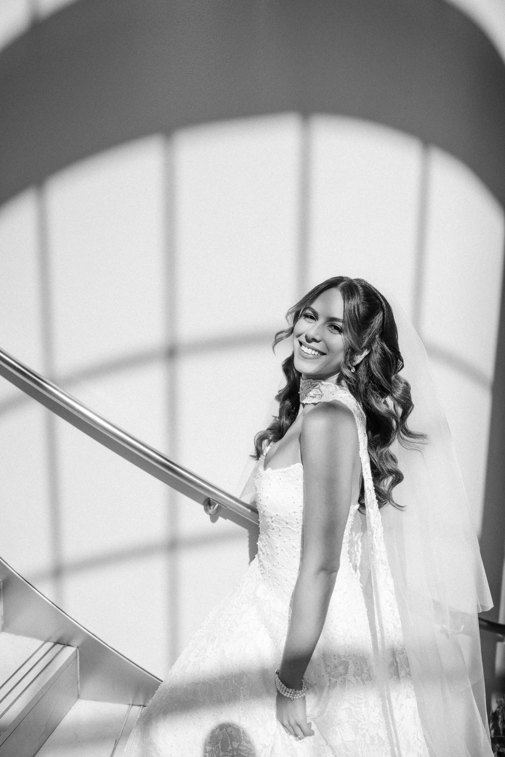 A bride in a white gown and veil stands on a staircase at Guastavino’s wedding, smiling at the camera, with sunlight casting shadows on the wall behind her.