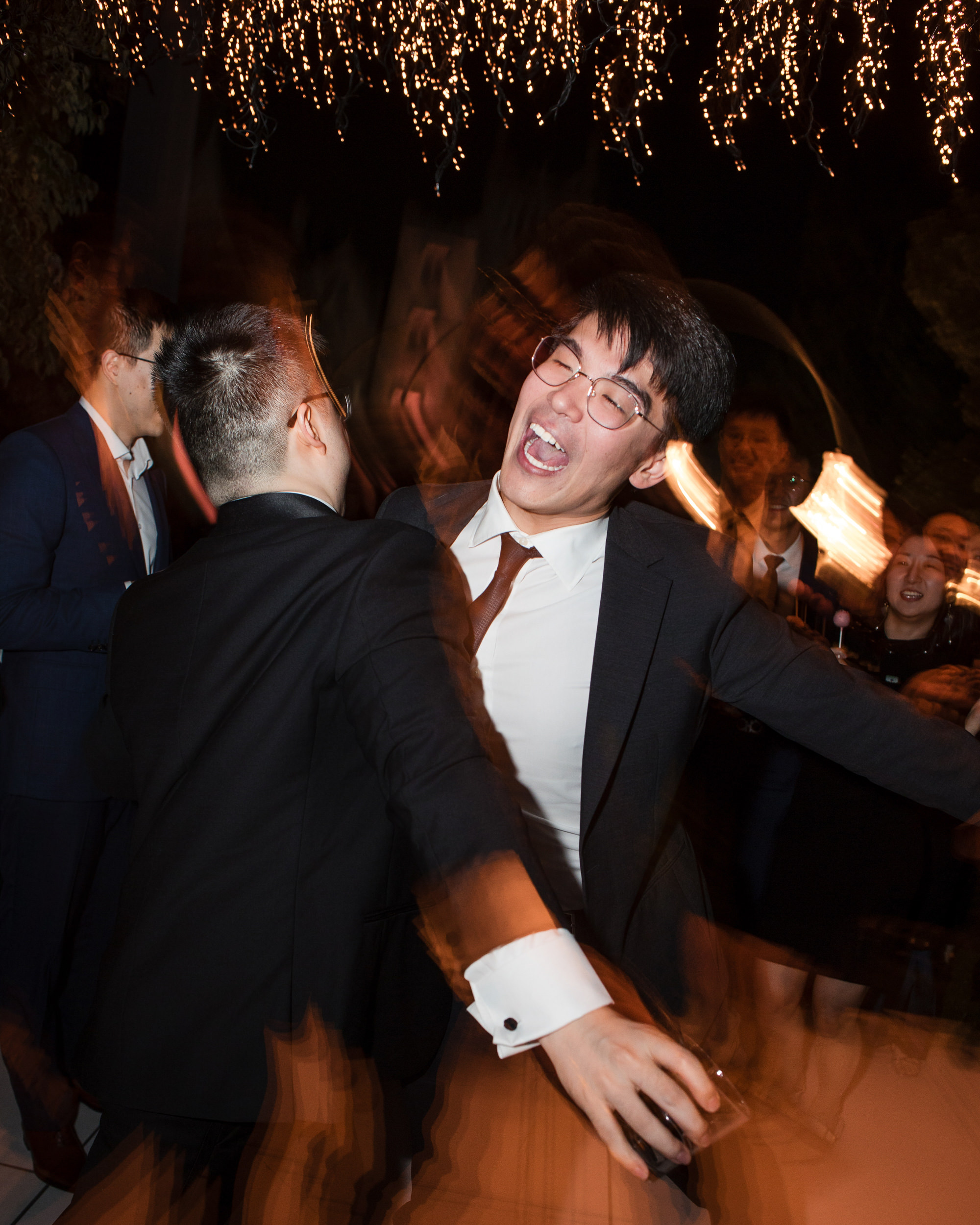 Two people in suits dance energetically at a nighttime event with string lights overhead at a Lyndhurst Mansion wedding; one appears mid-laugh while others look on in the background.